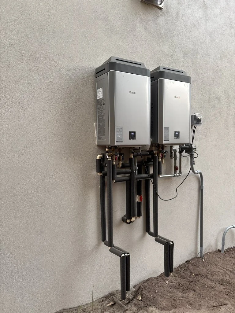 Two tankless water heaters mounted on an exterior wall with pipes and wiring.