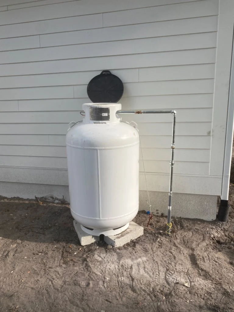 A white compressed air tank with a black lid mounted on a concrete block outside a building with white siding, connected via a metal pipe.