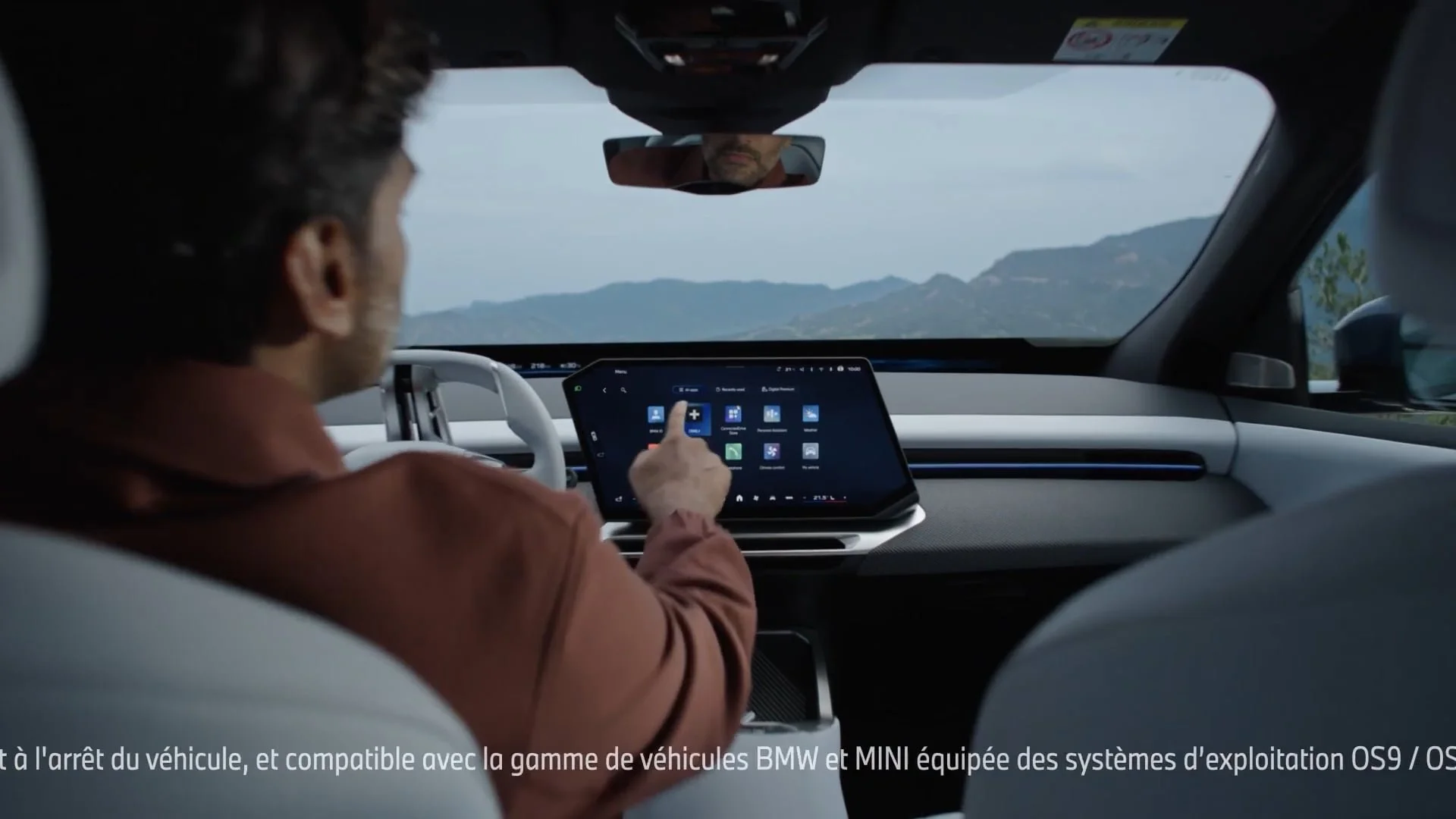 Un homme dans une voiture utilisant un écran tactile pour contrôler le système d'exploitation, avec vue sur un paysage montagneux à travers le pare-brise.