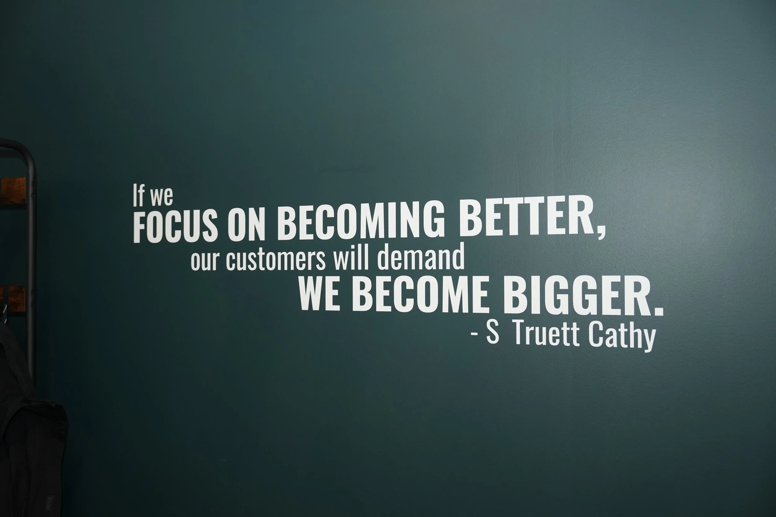 Part of a wall with a quote in white text that reads, "If we focus on becoming better, our customers will demand we become bigger." - S Truett Cathy. A corner of a black metal frame structure is visible on the left side of the image.