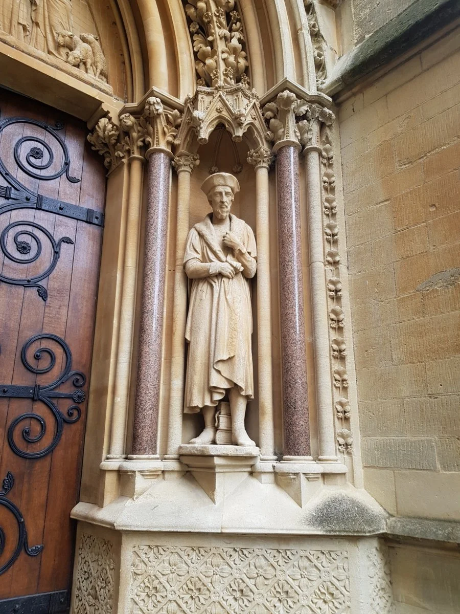 Stone sculpture of a man in historical attire, standing in an ornate architectural niche with columns and decorative elements, mounted on the wall near a wooden door.