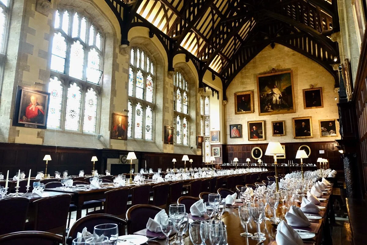 Elegant dining room with long tables set for a formal event, featuring white napkins, wine glasses, and candelabras. The room has high, arched stained glass windows and portraits on the walls, with a high vaulted ceiling supported by dark wooden beam