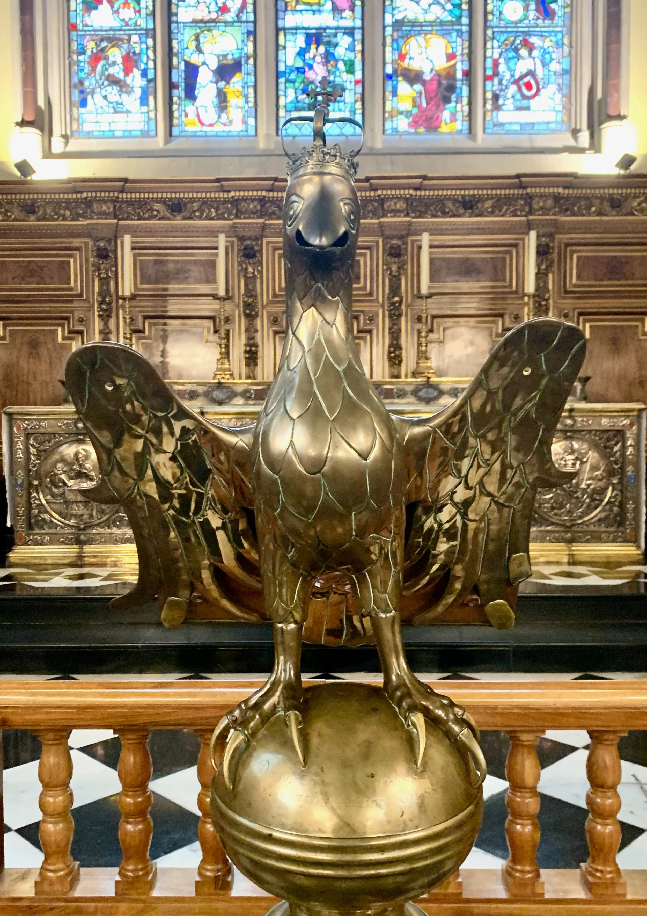A gold-colored sculpture of a phoenix or bird with its wings spread, positioned on a globe, inside a church or chapel with stained glass windows and ornate wooden decorations in the background.