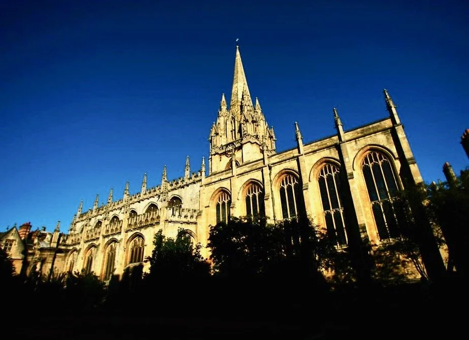 A Gothic-style cathedral with a tall central spire, large arched windows, and intricate stone details against a blue sky, surrounded by trees.