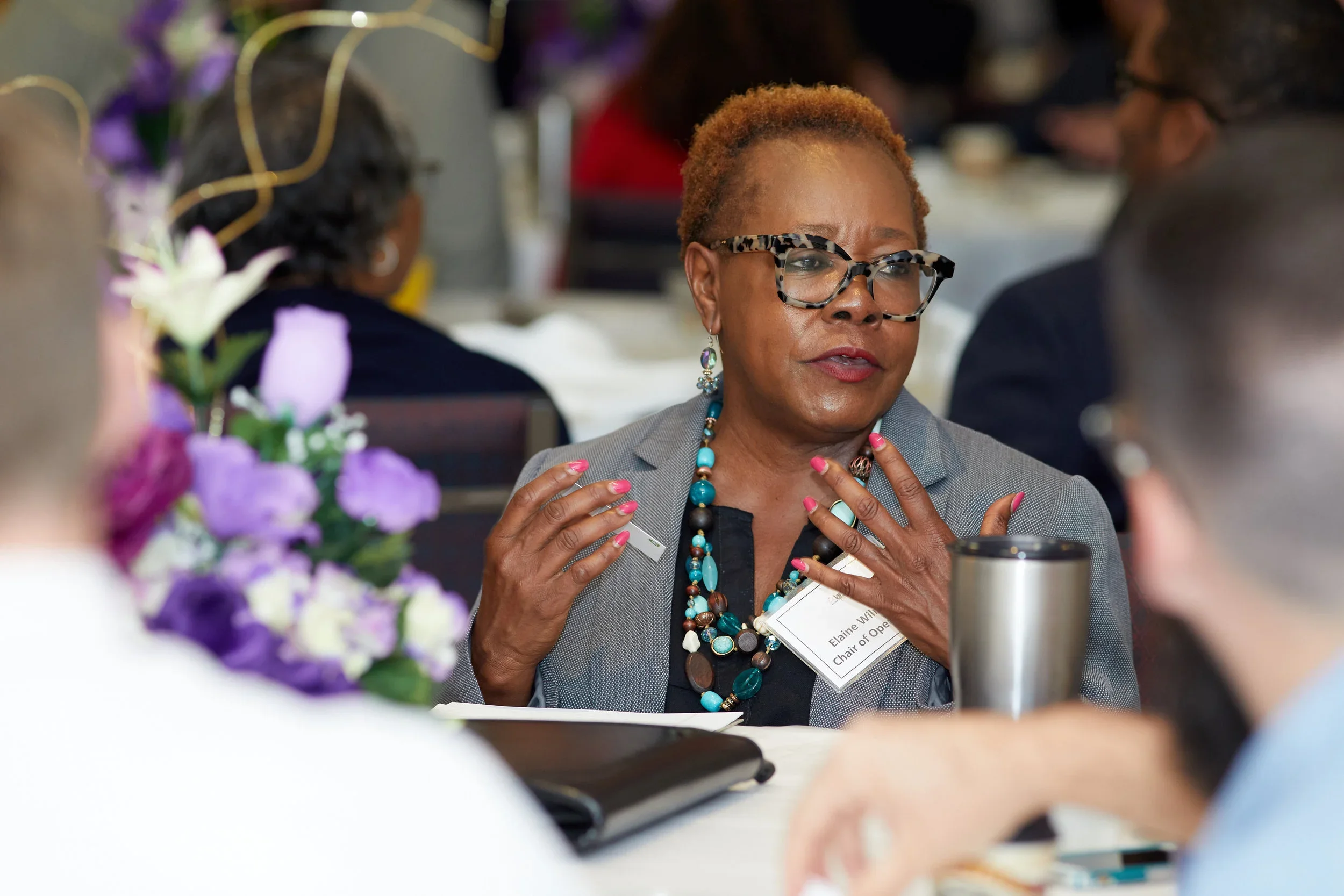 An older woman with short, reddish hair and glasses is speaking or explaining something at a meeting. She is wearing a gray blazer, large beaded necklace, earrings, and has a name tag that reads 'Elaine W...'.