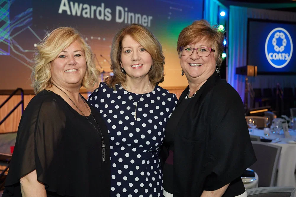 Three women standing together at an awards dinner, smiling, in a banquet hall with tables set for a formal event.