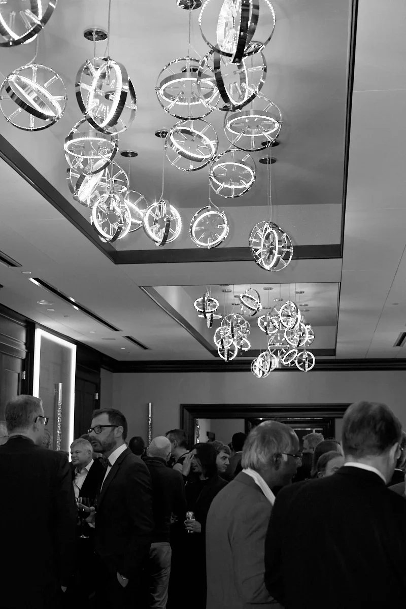 A black and white photo of a crowded indoor event with many people dressed in formal attire. The ceiling features modern chandelier light fixtures resembling rings or circles.