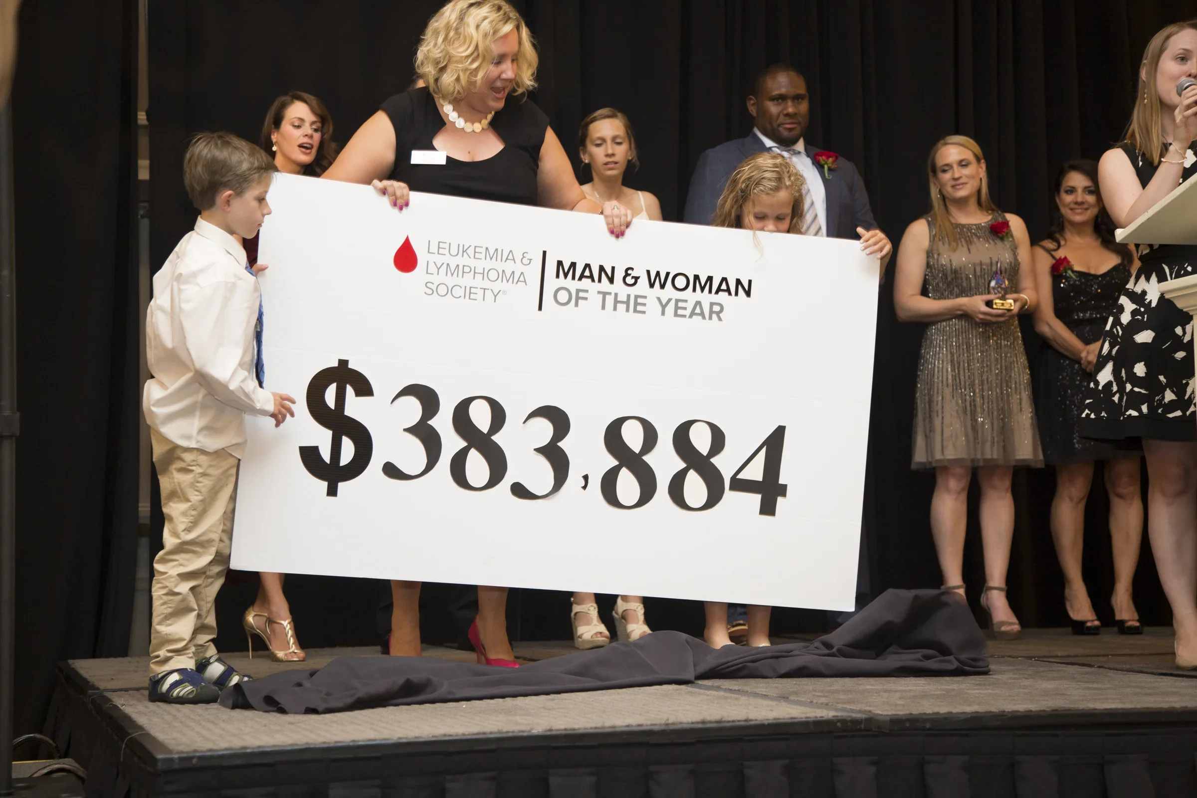 People on stage holding a giant check for $383,884 at the Leukemia & Lymphoma Society event, with a woman speaking into a microphone.