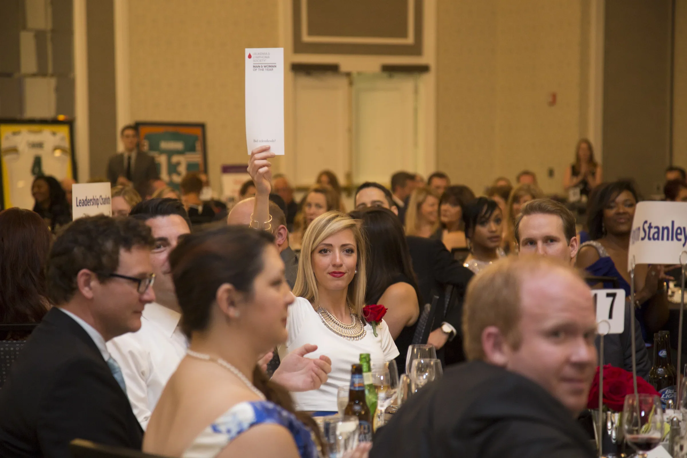 A diverse group of people seated at a formal event or banquet, some looking towards the camera, with table signs and drinks visible on the table.