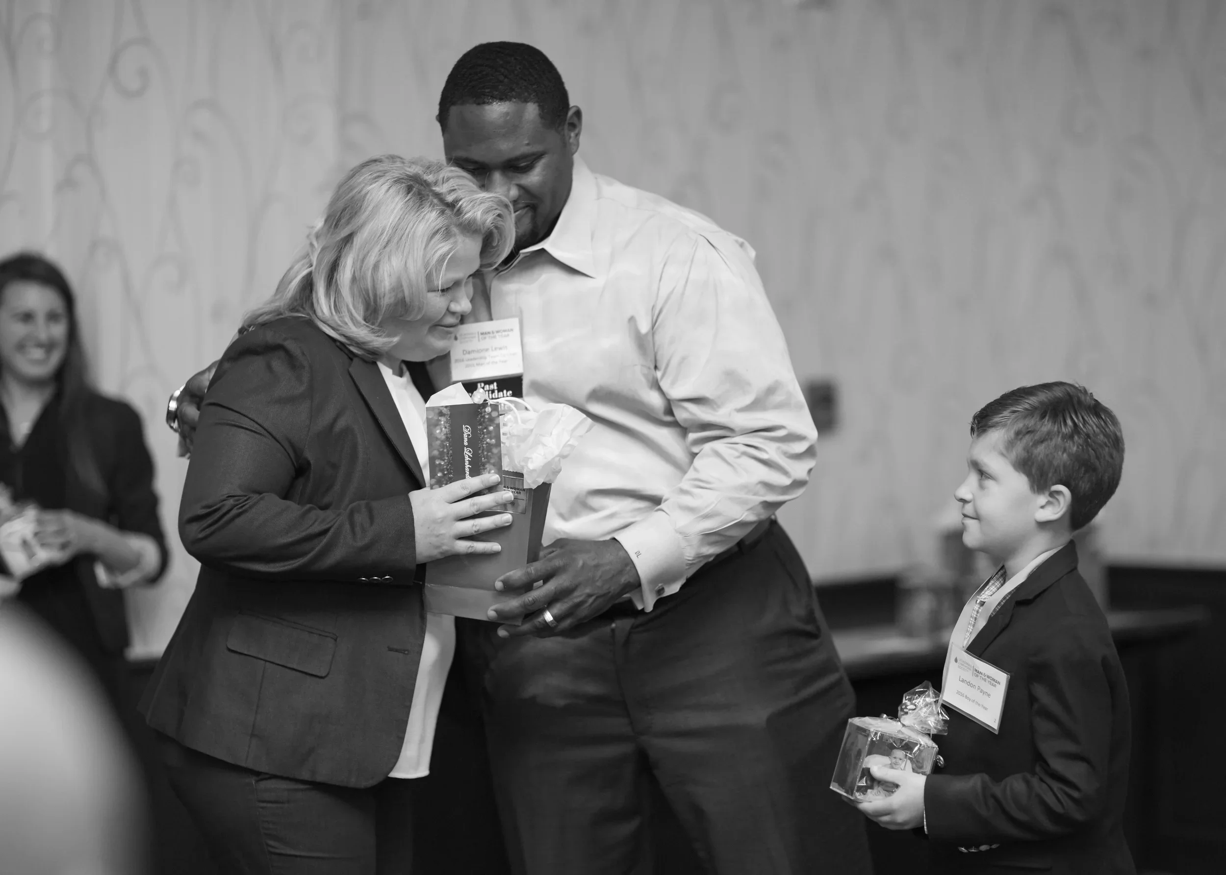 A woman and a tall man sharing a moment during an award or recognition event. The woman is holding a gift bag, smiling, while the man is helping with the bag. A young boy in a suit and tie, holding a small gift, is looking up at them.