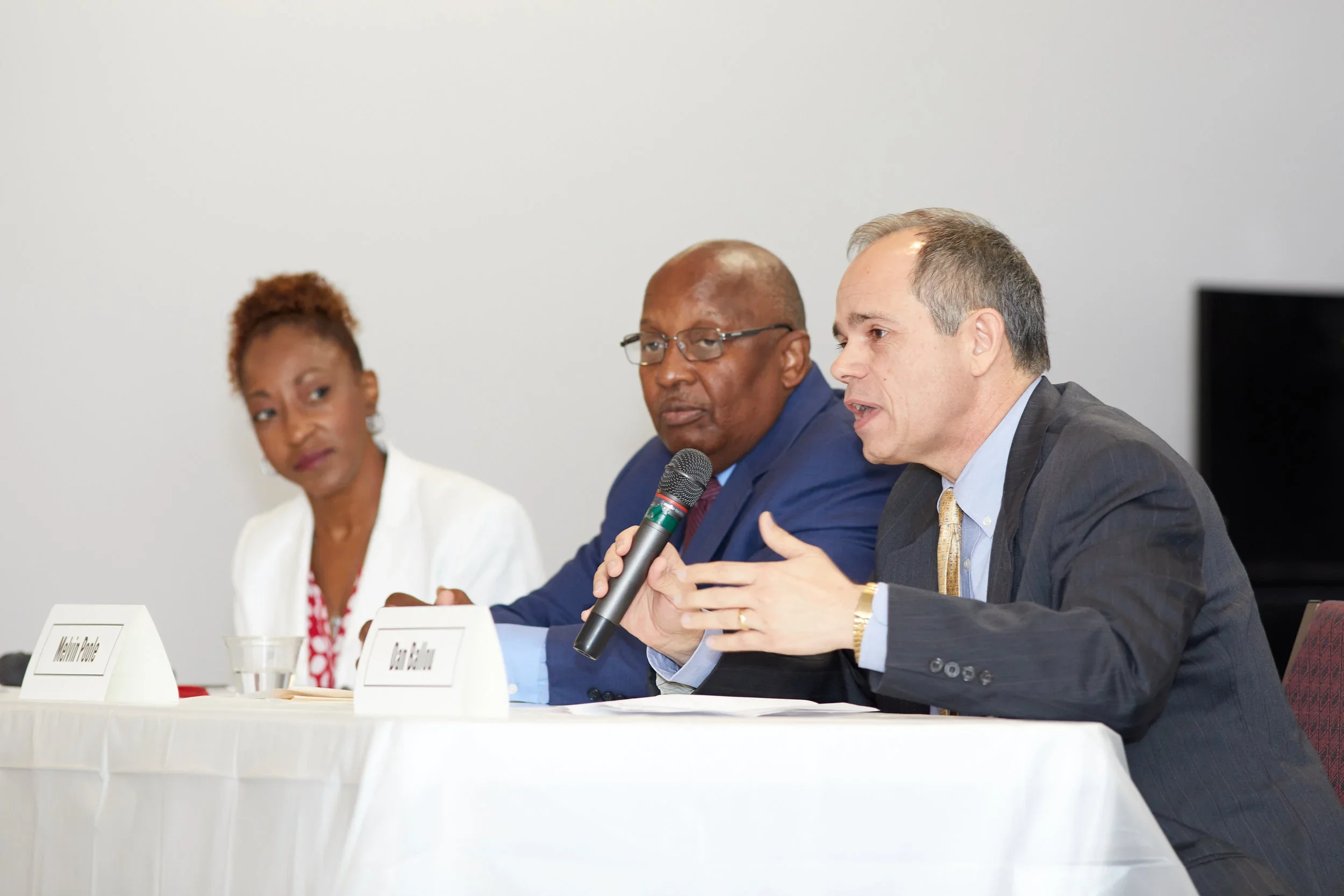 Three people sitting at a panel discussion table, with white name tags, in a conference room. The man on the right is speaking into a microphone, wearing a dark suit, light shirt, and gold watch. The man in the middle, wearing glasses and a blue suit