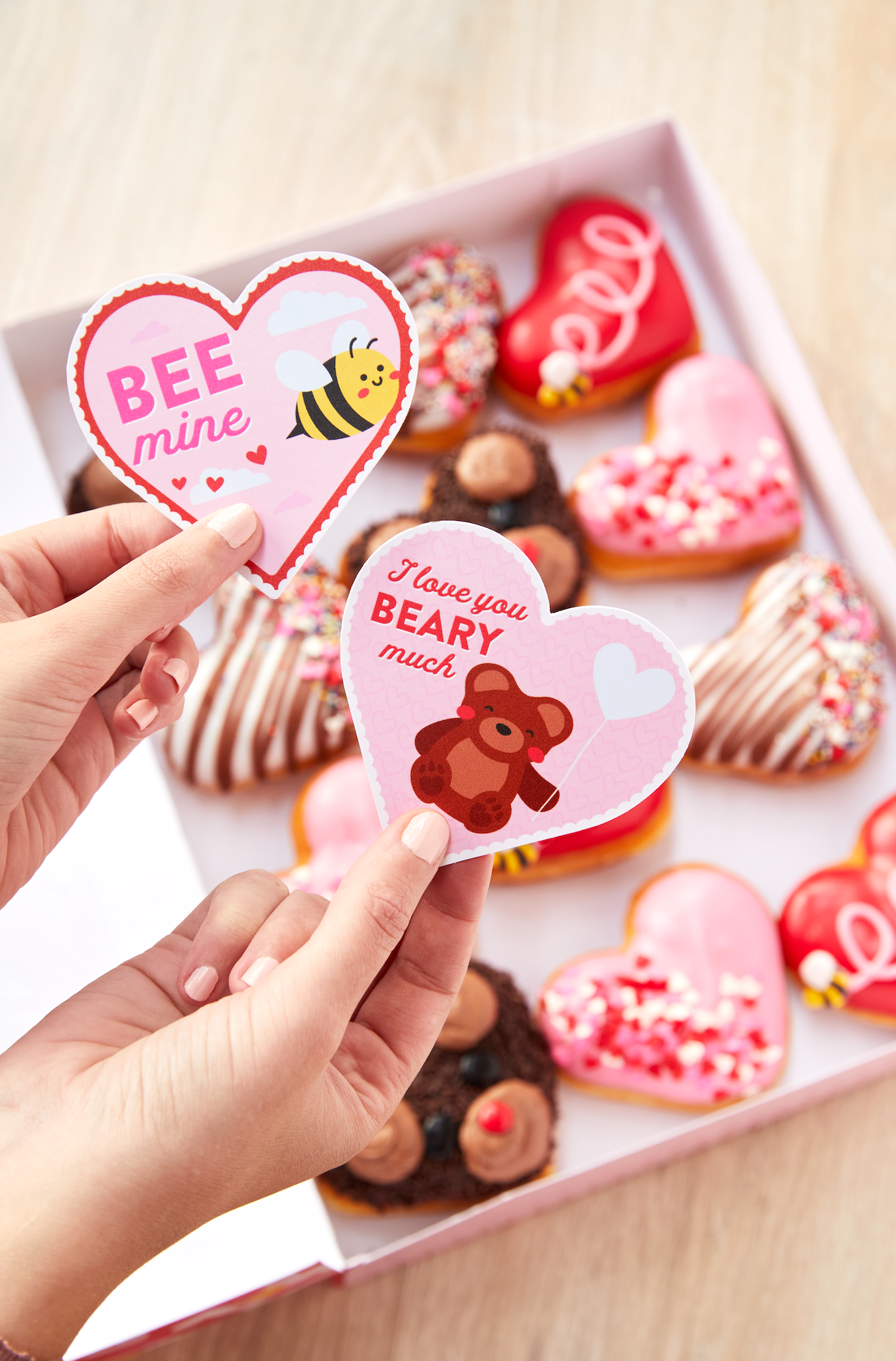 Hands holding heart-shaped Valentine's cards with cute designs, above a box of assorted heart-shaped donuts with colorful toppings.