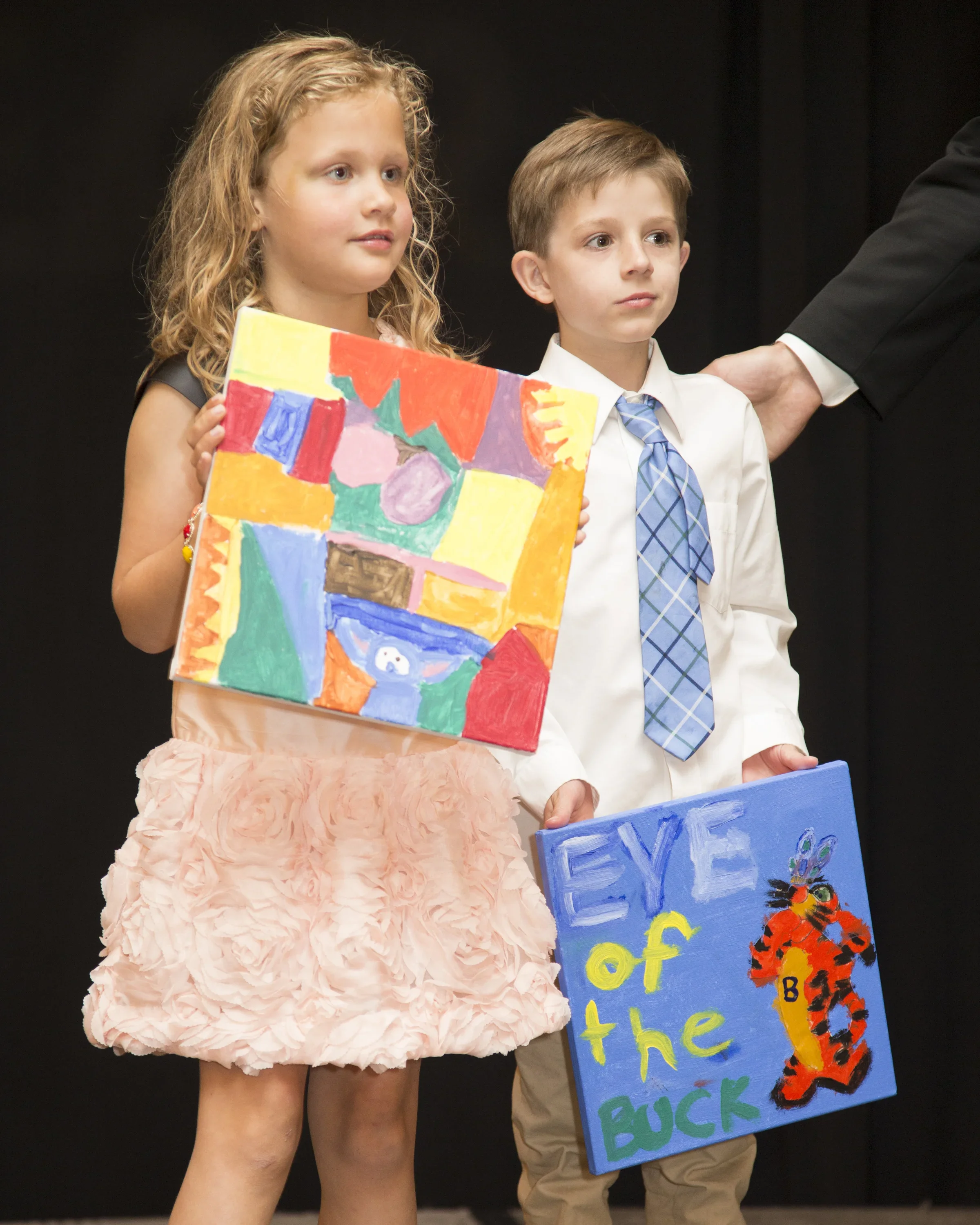 Two children, a girl with curly hair wearing a pink ruffled skirt and a boy with short hair in a white shirt and blue tie, are standing on stage holding colorful signs. The girl’s sign has abstract artwork with various bright colors, and the boy’s si