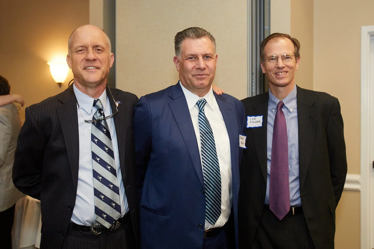 Three men in suits standing side by side at an indoor event, smiling for the camera.