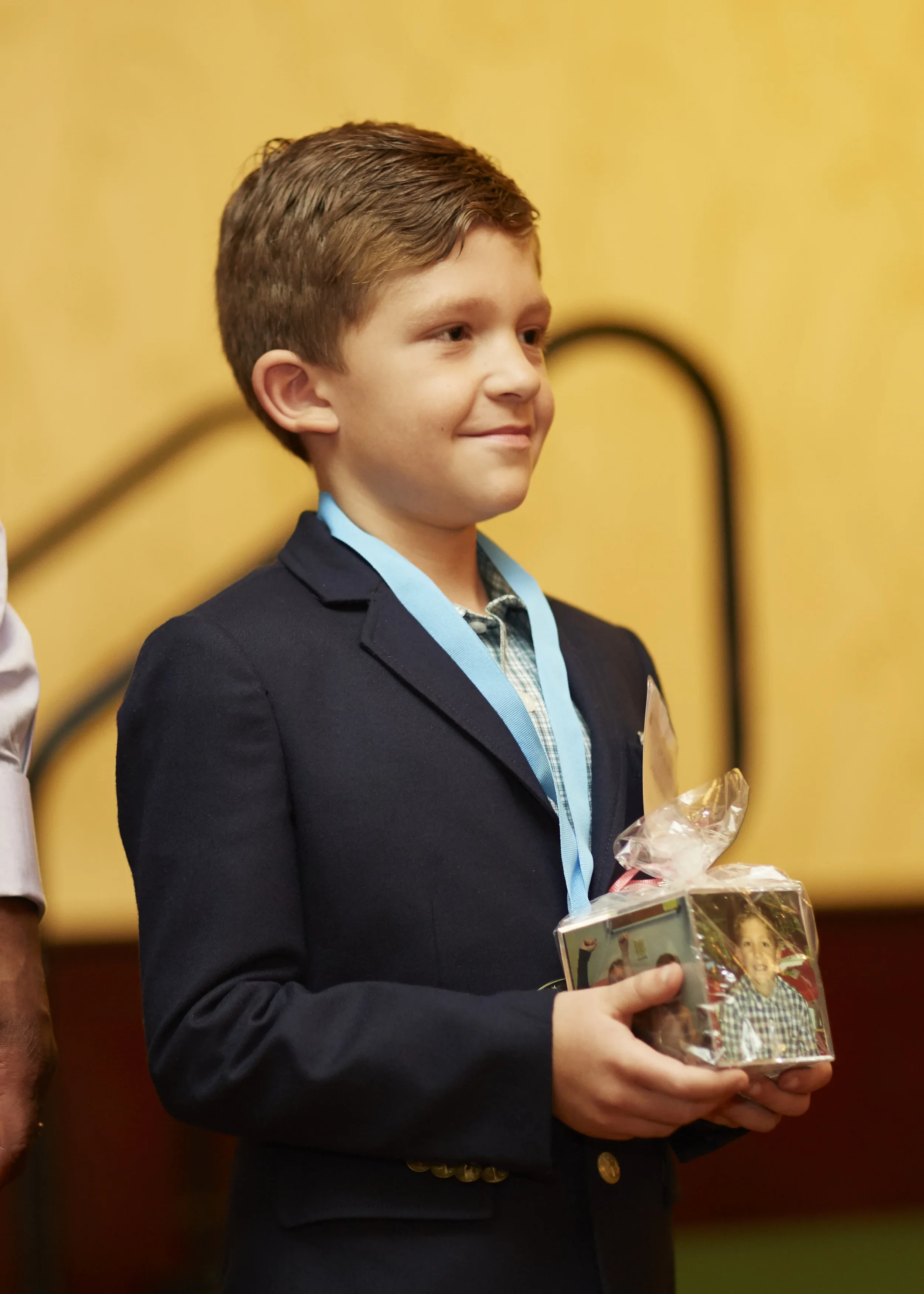 Young boy in a dark blazer holding a wrapped gift with a photo of himself on it, smiling at an award ceremony.