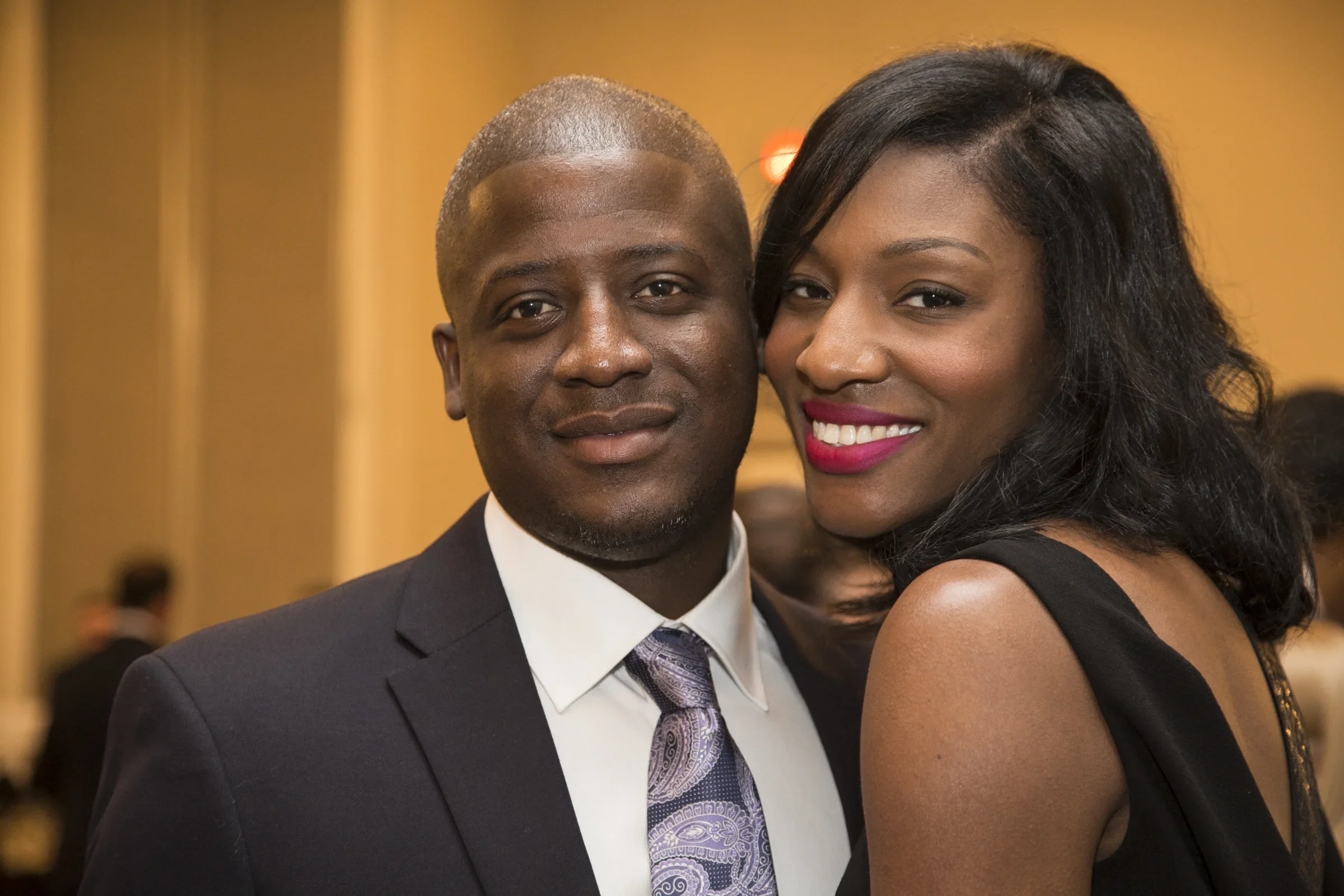 A man and woman posing closely for a photo indoors, both dressed in formal attire. The man is wearing a suit, shirt, and tie, and the woman is wearing a sleeveless black dress with curly black hair and bright lipstick.