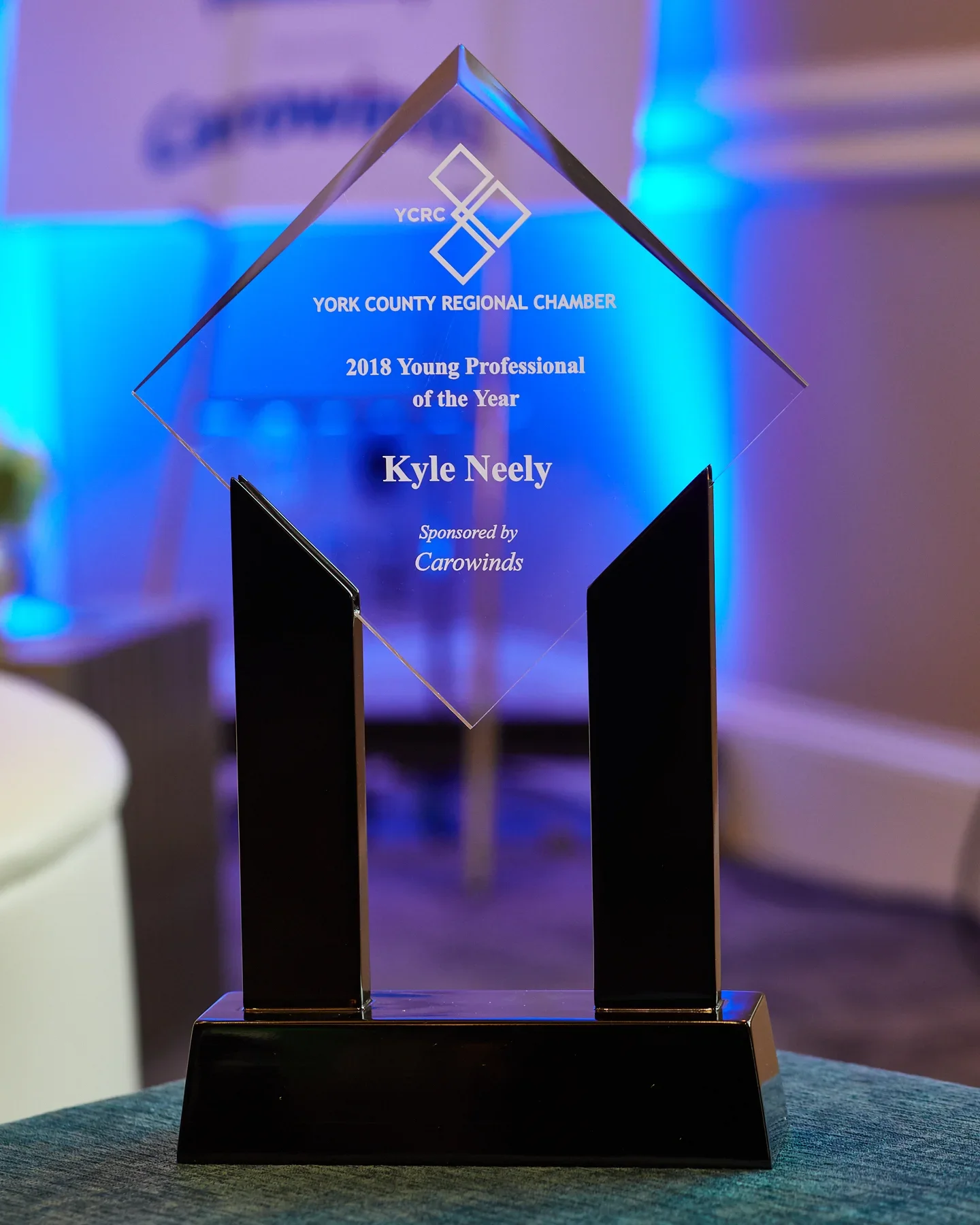 Glass award plaque with black base for 2018 Young Professional of the Year, issued by York County Regional Chamber and sponsored by Carowinds, with blue and purple lighting in the background.