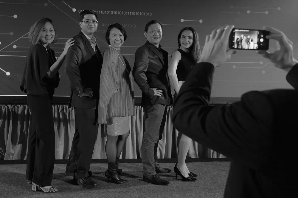 Group of five diverse professionals posing for a photo on stage, with someone taking their picture using a smartphone, in a conference or event setting.