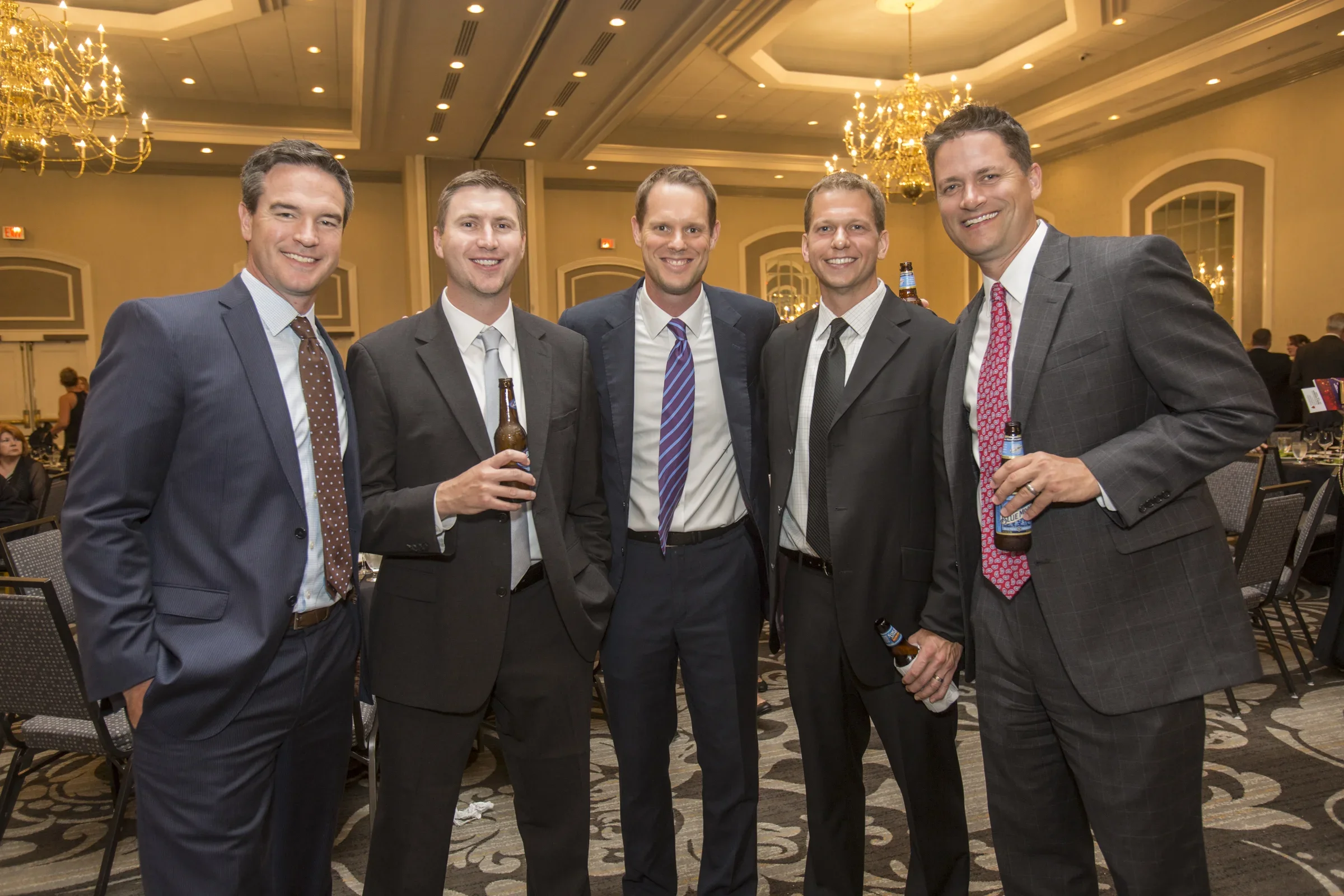 Five men in suits at a formal event, standing together and smiling, holding beer bottles, in a decorated banquet hall with chandeliers.