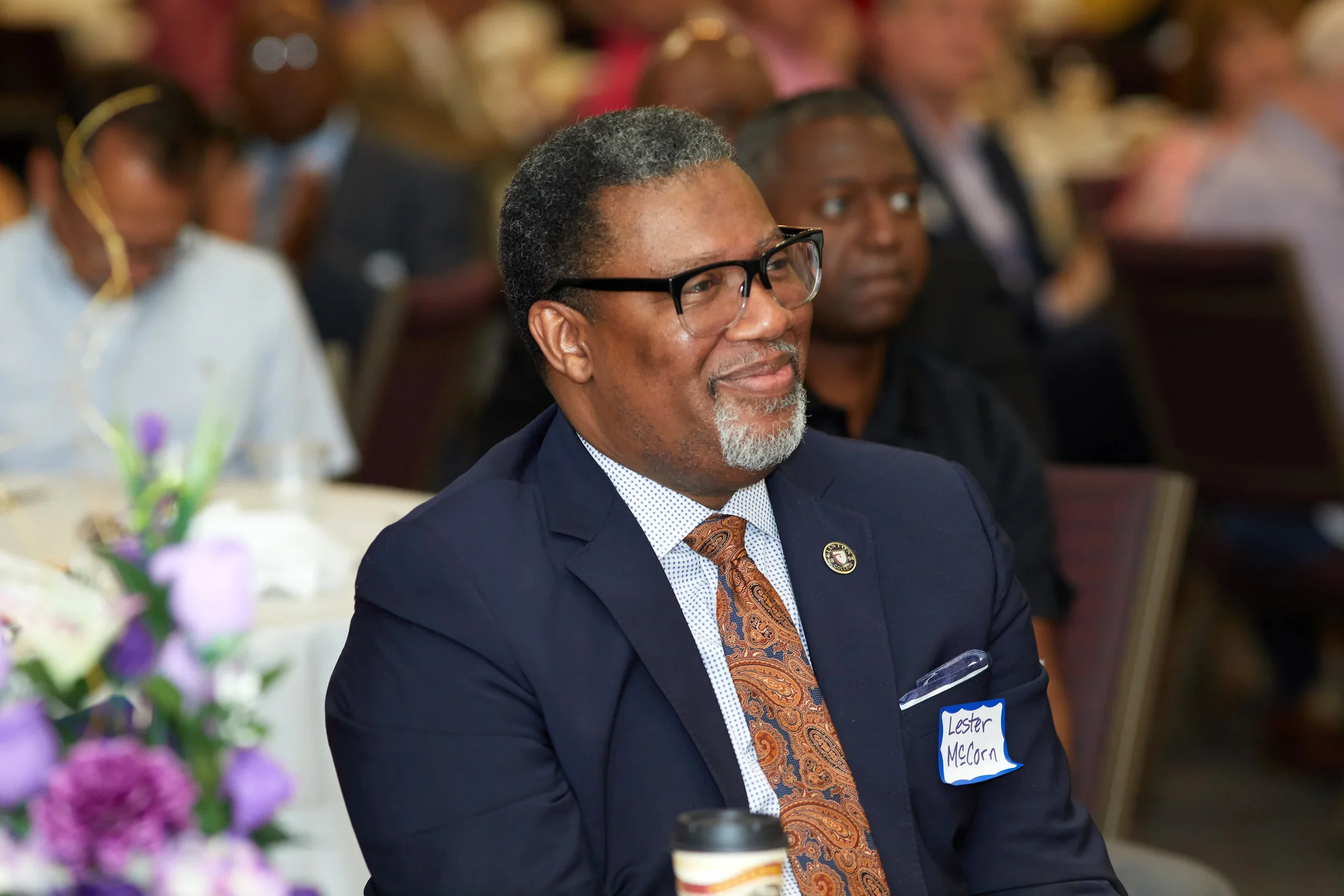 A man wearing glasses and a navy suit sits sideways at what looks like a banquet table looking off screen perhaps at a stage. 