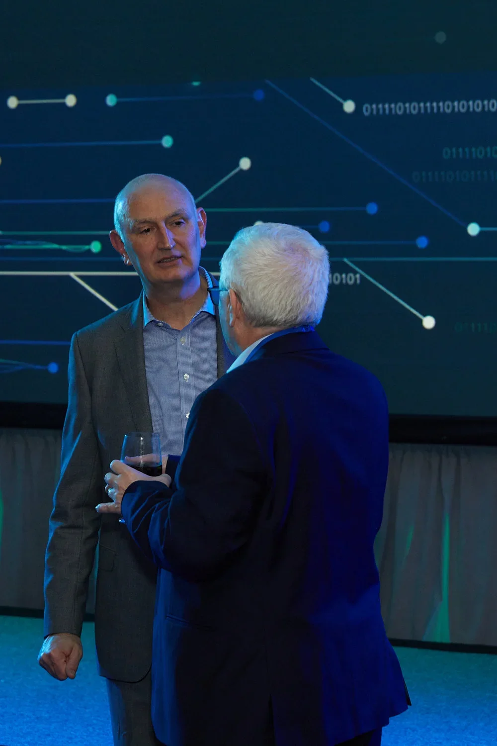 Two men in suits having a conversation at a technology conference. One is holding a glass of red wine, and there is a digital screen with binary numbers and data lines in the background.