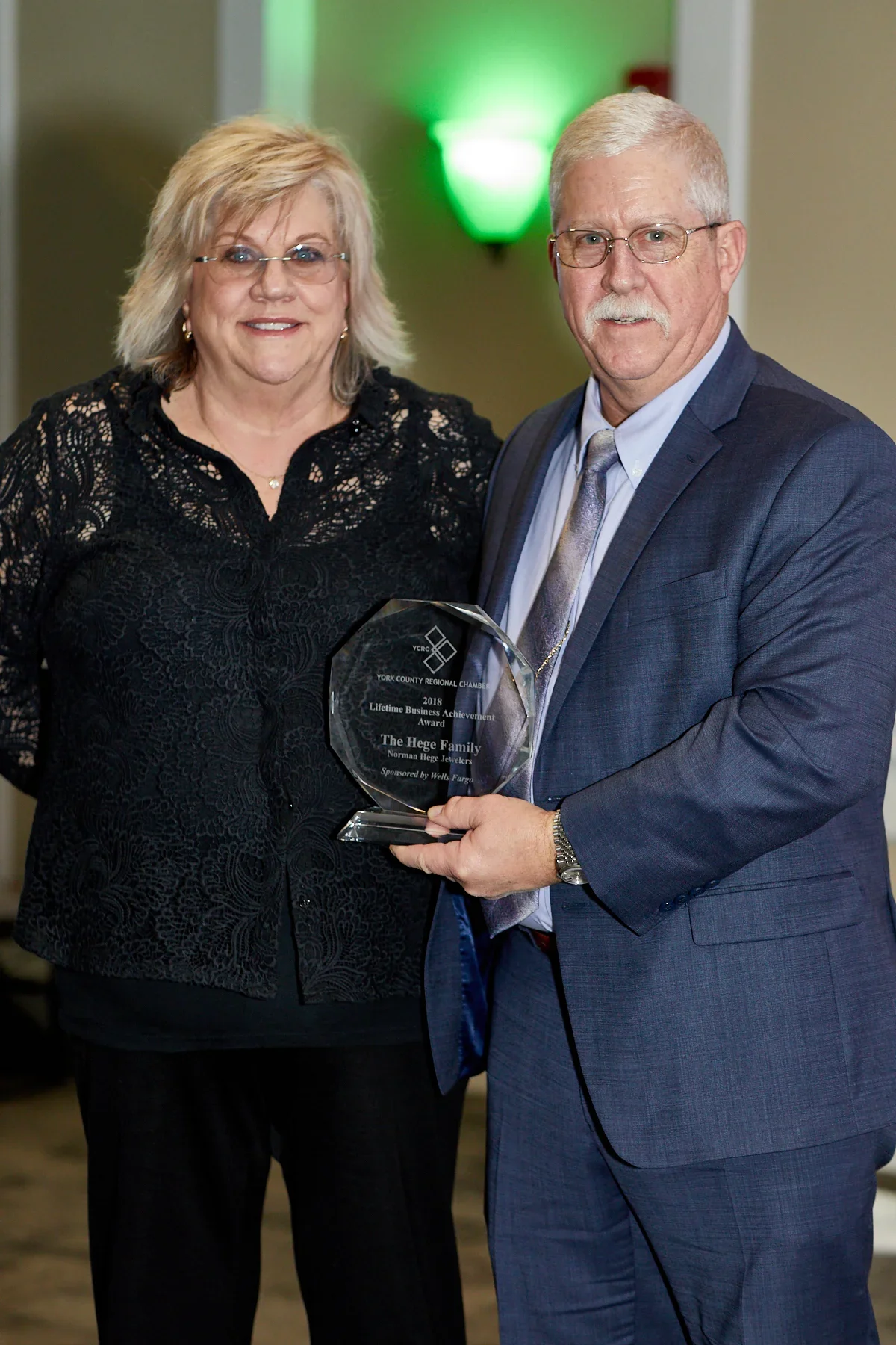A man in a blue suit and glasses holding a glass award plaque, standing next to a woman with blonde hair wearing a black lace blouse, both smiling at the camera during an award ceremony.
