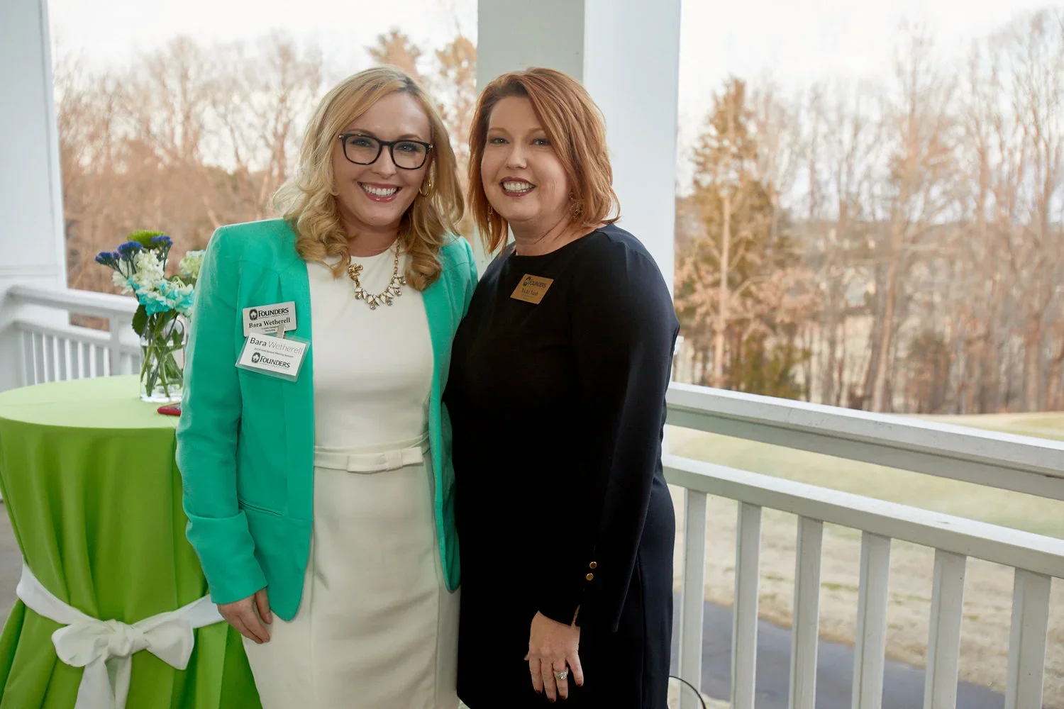 Two women stand close together on a porch with a green table and flowers in the background. The woman on the left wears glasses, a turquoise blazer, and a white dress, with name tags that say 'Bara Wetherell.' The woman on the right wears a black dre