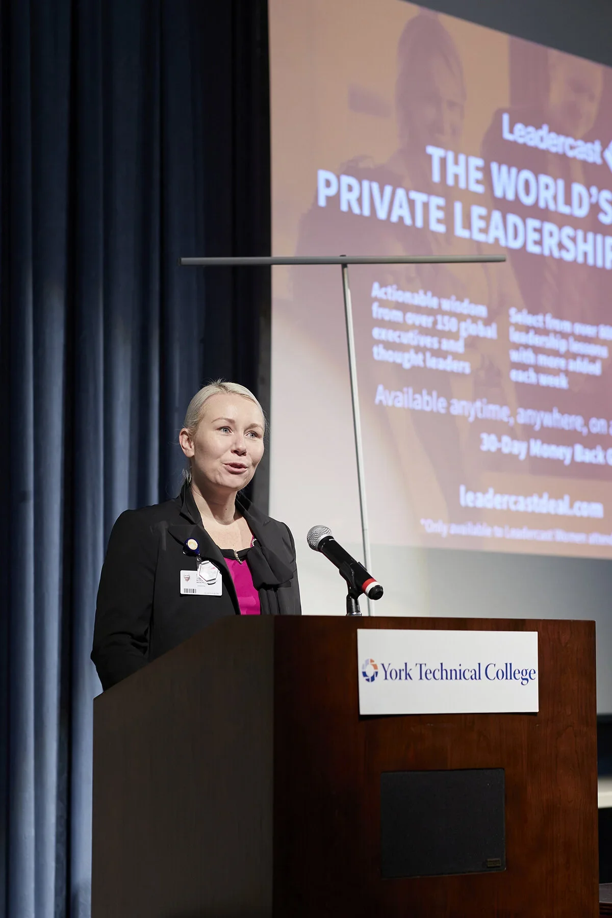 A woman in business attire speaking at a podium with a York Technical College logo, with a large screen behind her displaying a presentation about private leadership.