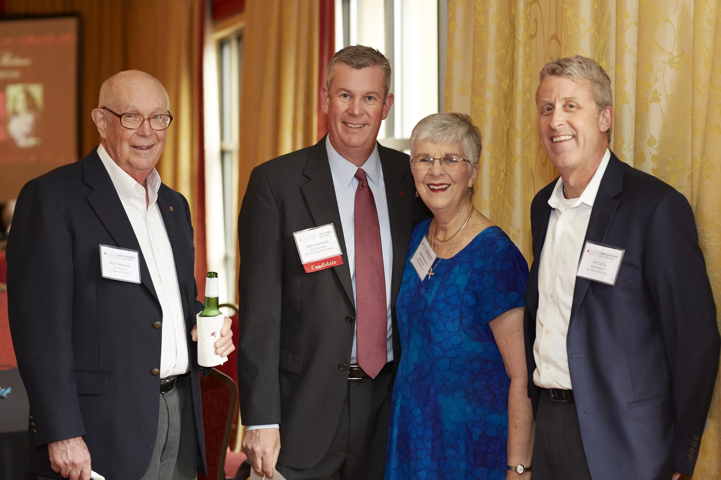 Four people, two men and a woman between them, smiling and posing for a photo at a formal event indoors, with yellow curtains and a red and yellow backdrop in the background.