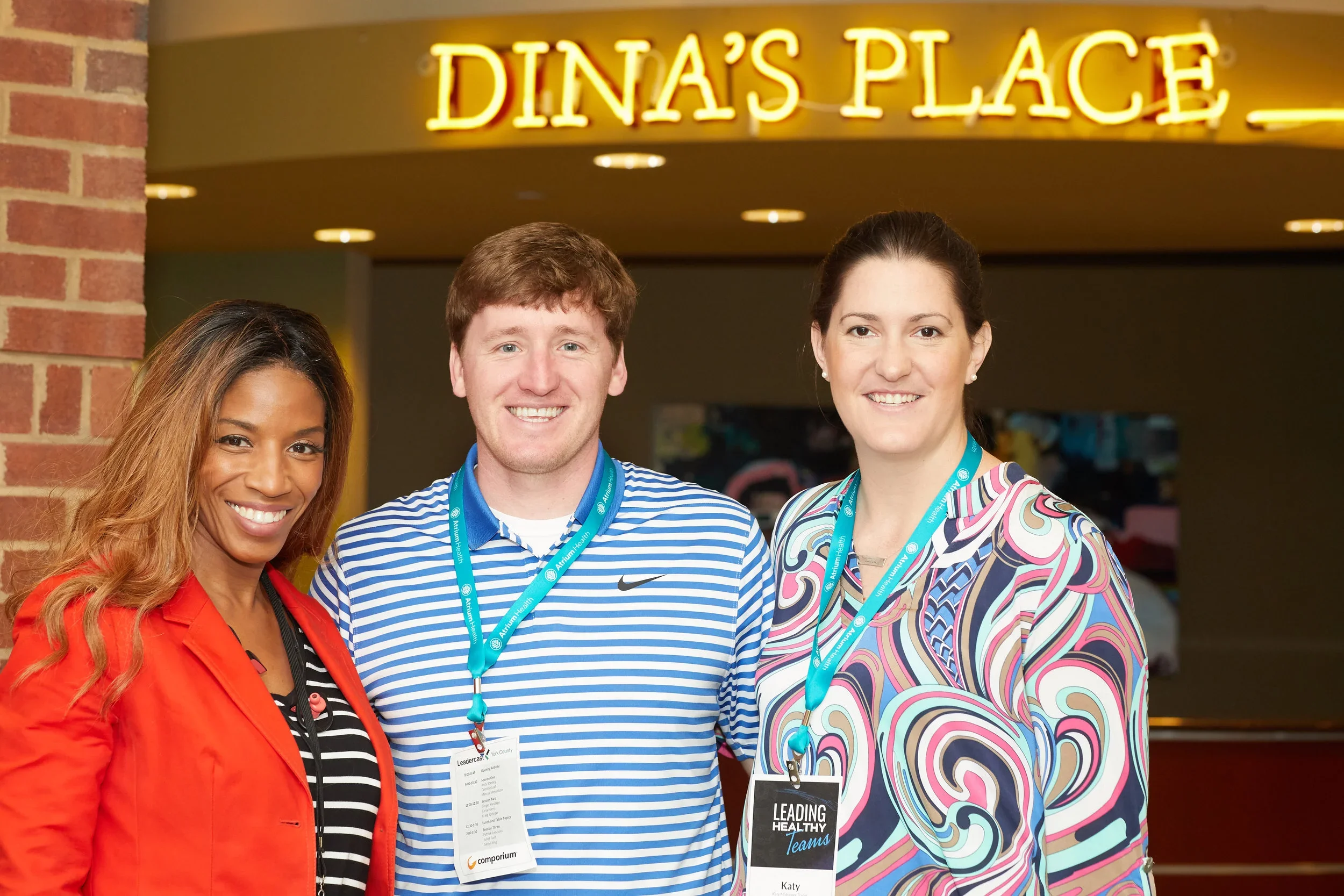 Three individuals smiling at a conference or event standing in front of a sign that says DINA'S PLACE, wearing conference badges and lanyards.
