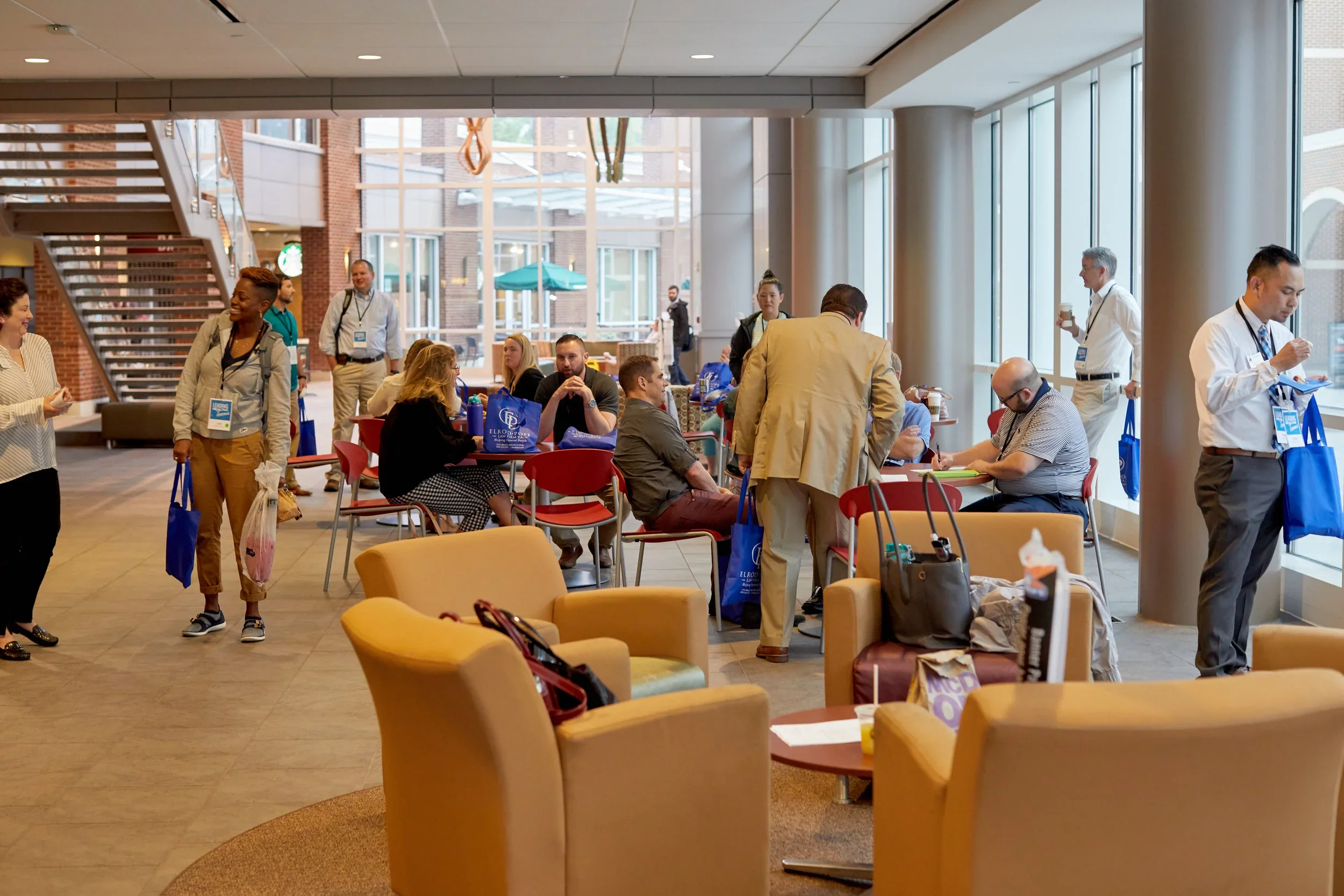 People gathered in a bright, modern indoor space with large windows, some sitting at tables, others standing. The room has a staircase, chairs, and tables with bags and papers.