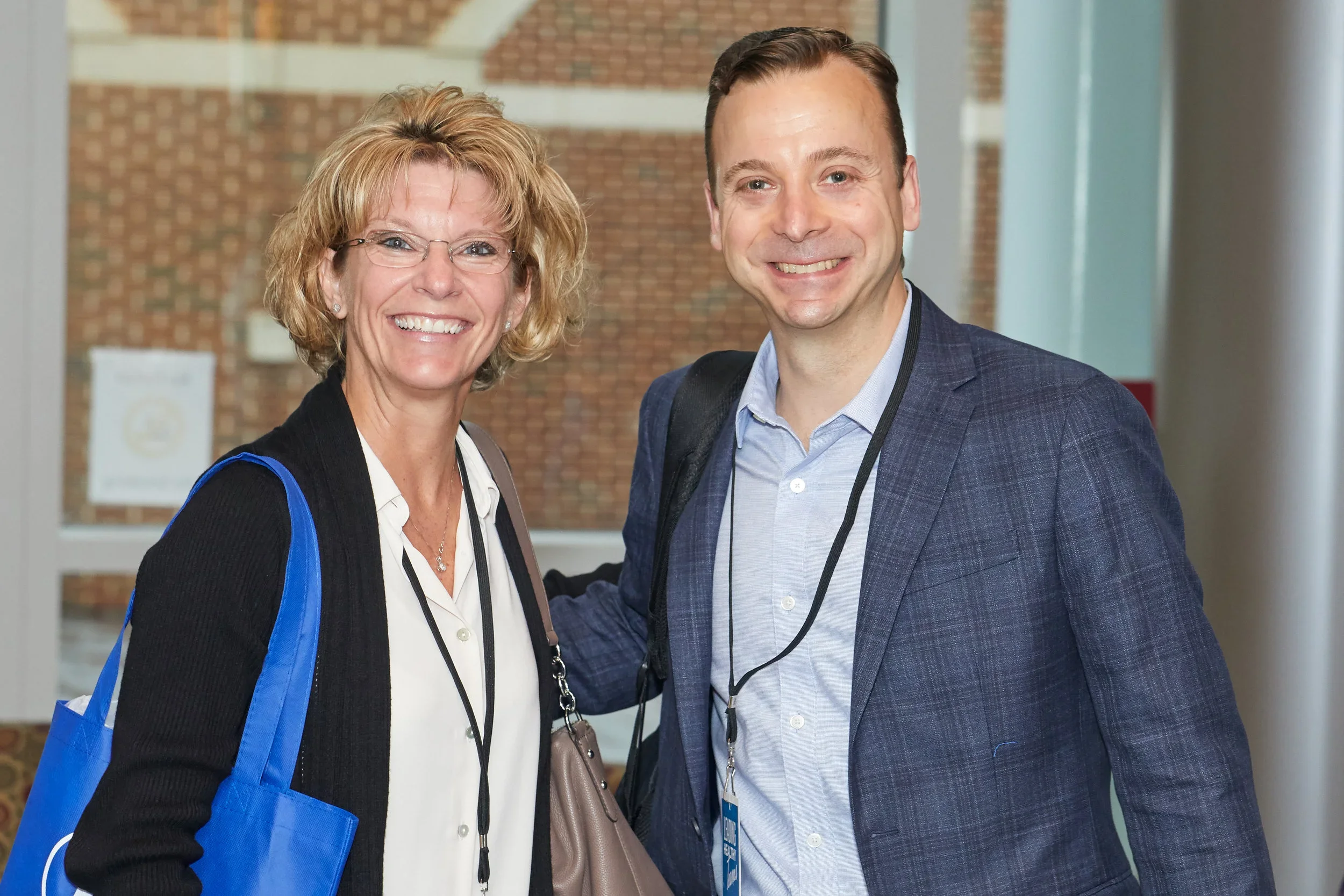 A woman and man smiling at a conference, with name tags and conference lanyards, in an indoor setting with brick wall background.