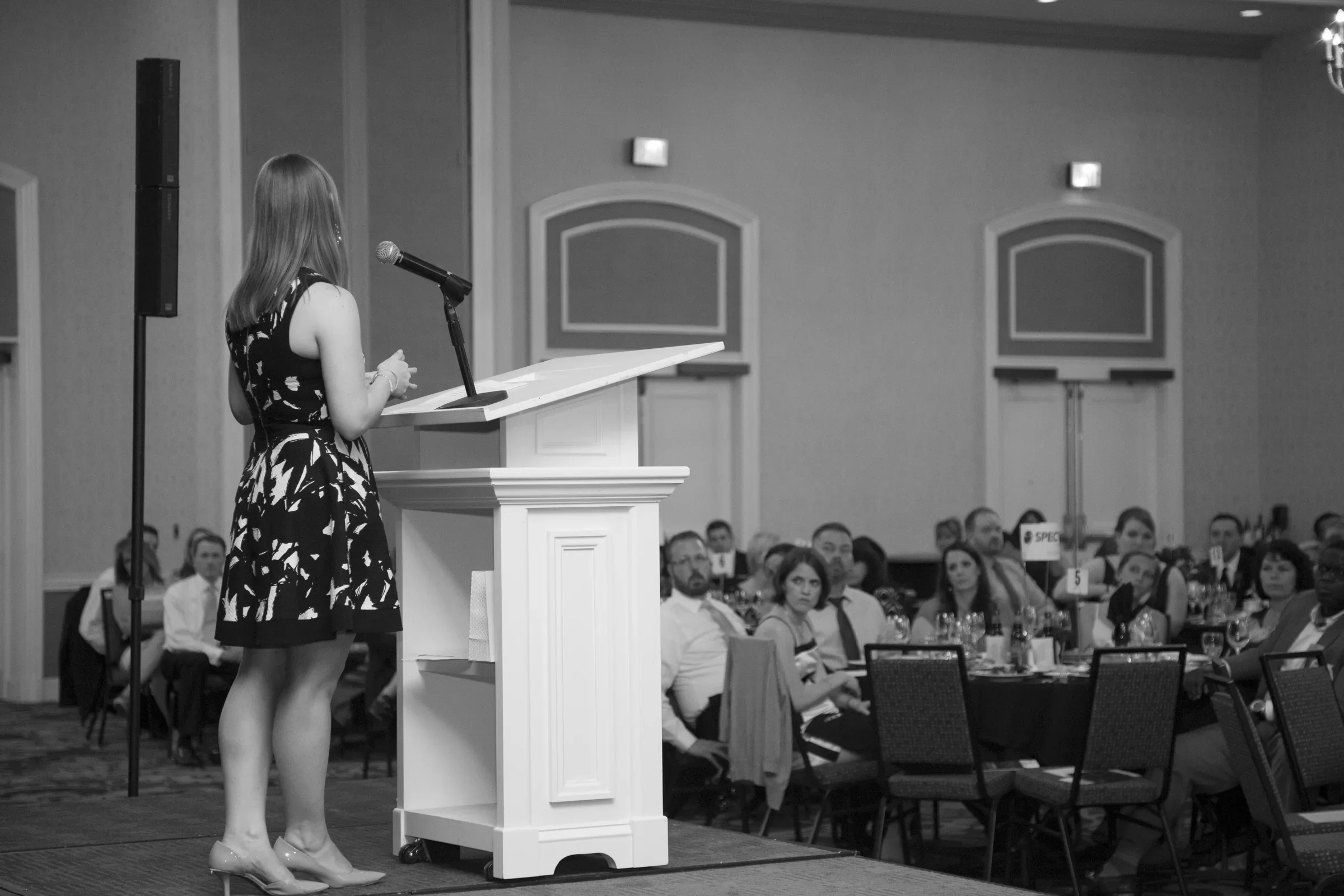 A woman giving a speech at a podium in a conference room, with an audience seated at tables.