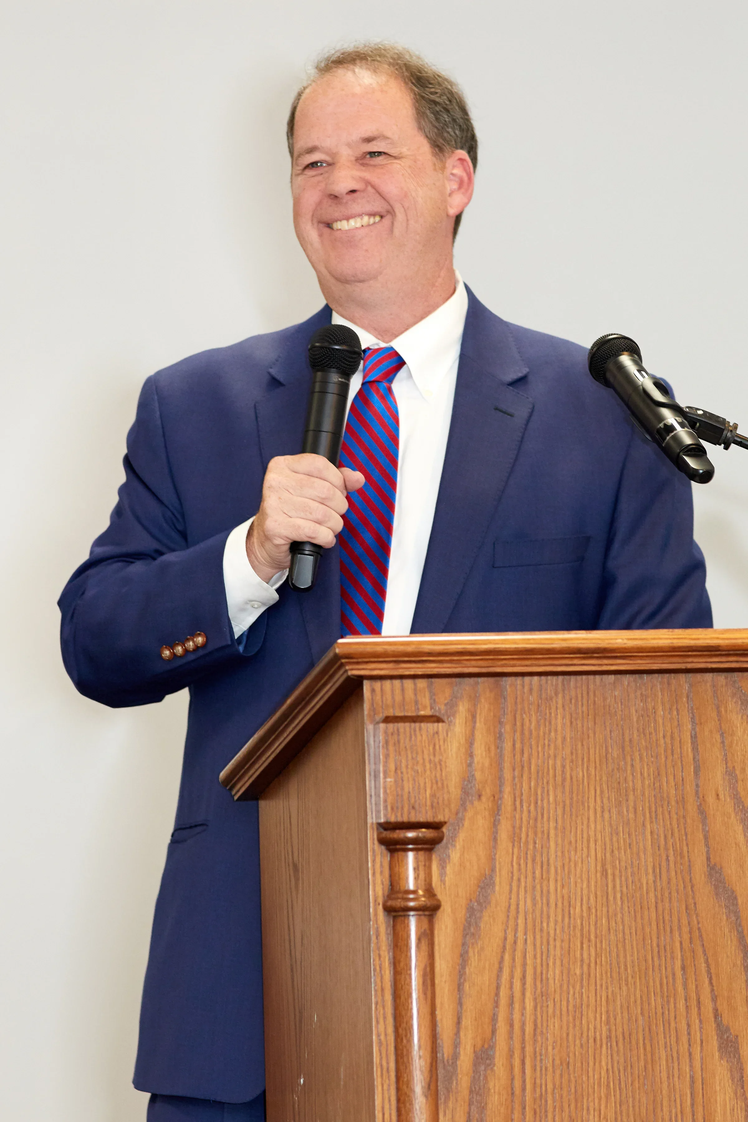 Man in a blue suit giving a speech at a wooden podium, holding a microphone, smiling, with another microphone attached to the podium.