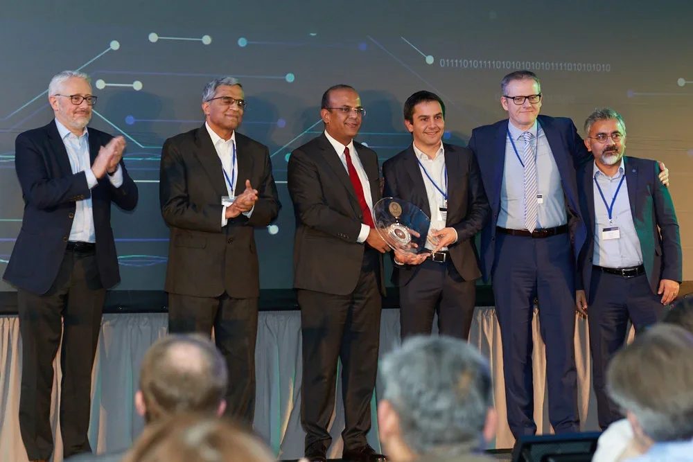 Group of six men in formal suits on stage during award ceremony, with one man holding a clear award trophy. Audience members are seated in front. Digital graphics and binary code are projected on screen behind them.