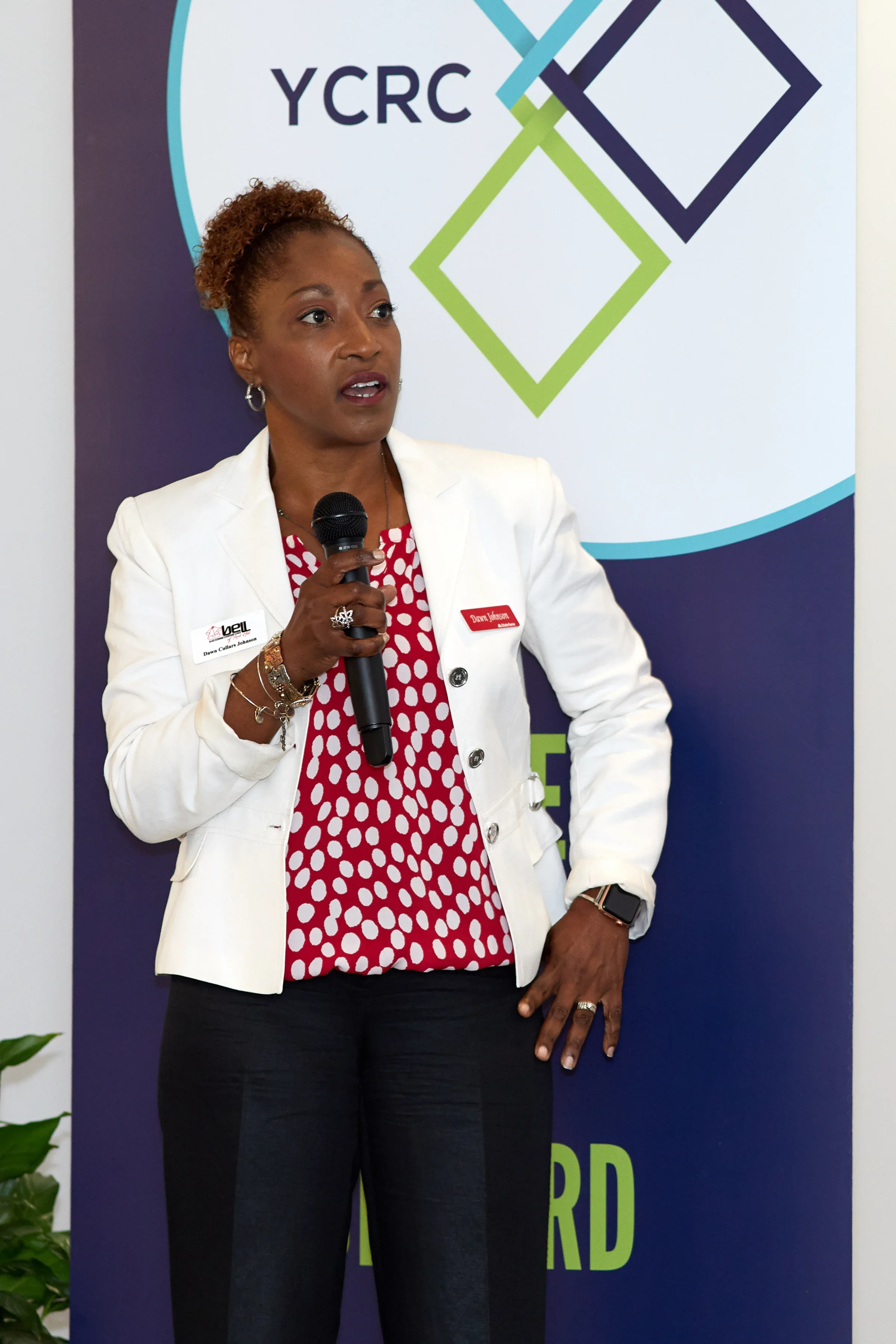 A woman in a white blazer holding a microphone, standing in front of a backdrop with the YCRC logo and geometric design.