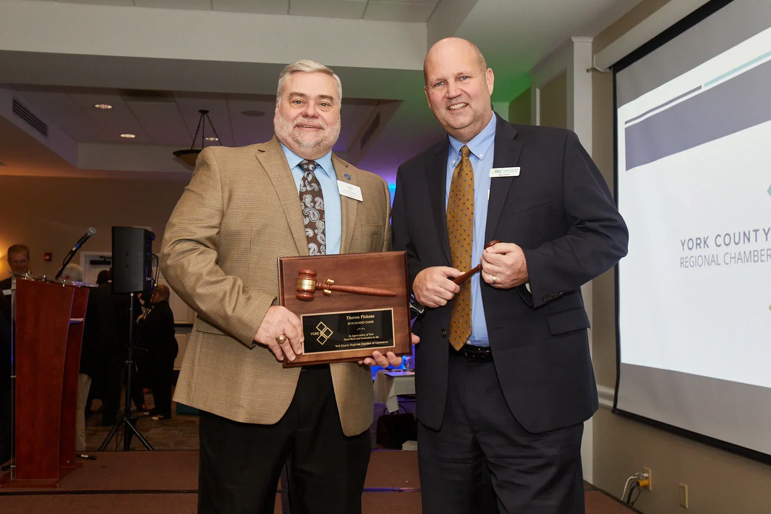 Two men in suits standing together at an award ceremony, one holding a plaque with a gavel on it, in a conference room with a projector screen and microphones.