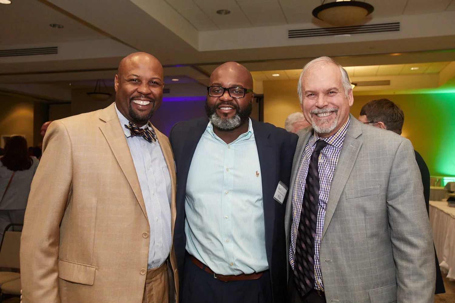 Three men in business attire smiling at an indoor event with others in the background.