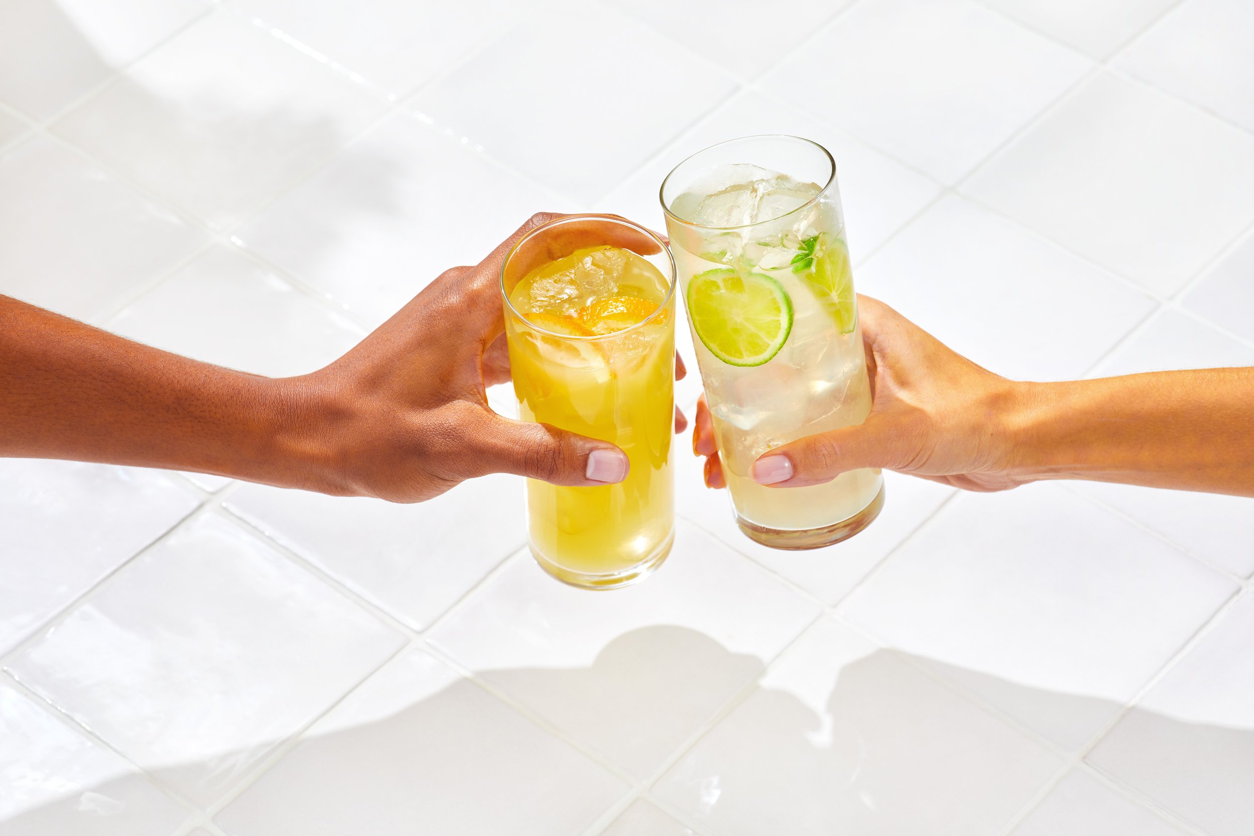 Two hands toasting with alcoholic beverages; one with a yellow drink and the other with a drink containing lime slices on a white tiled background.