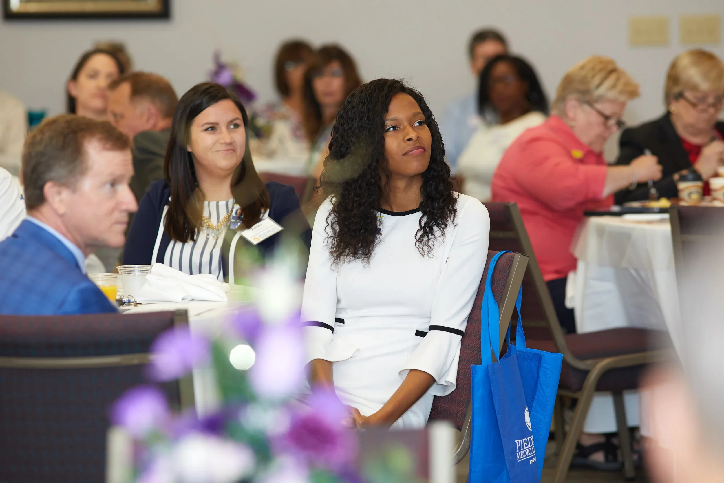 A diverse group of women and men attending a conference or seminar, sitting at round tables, listening attentively.