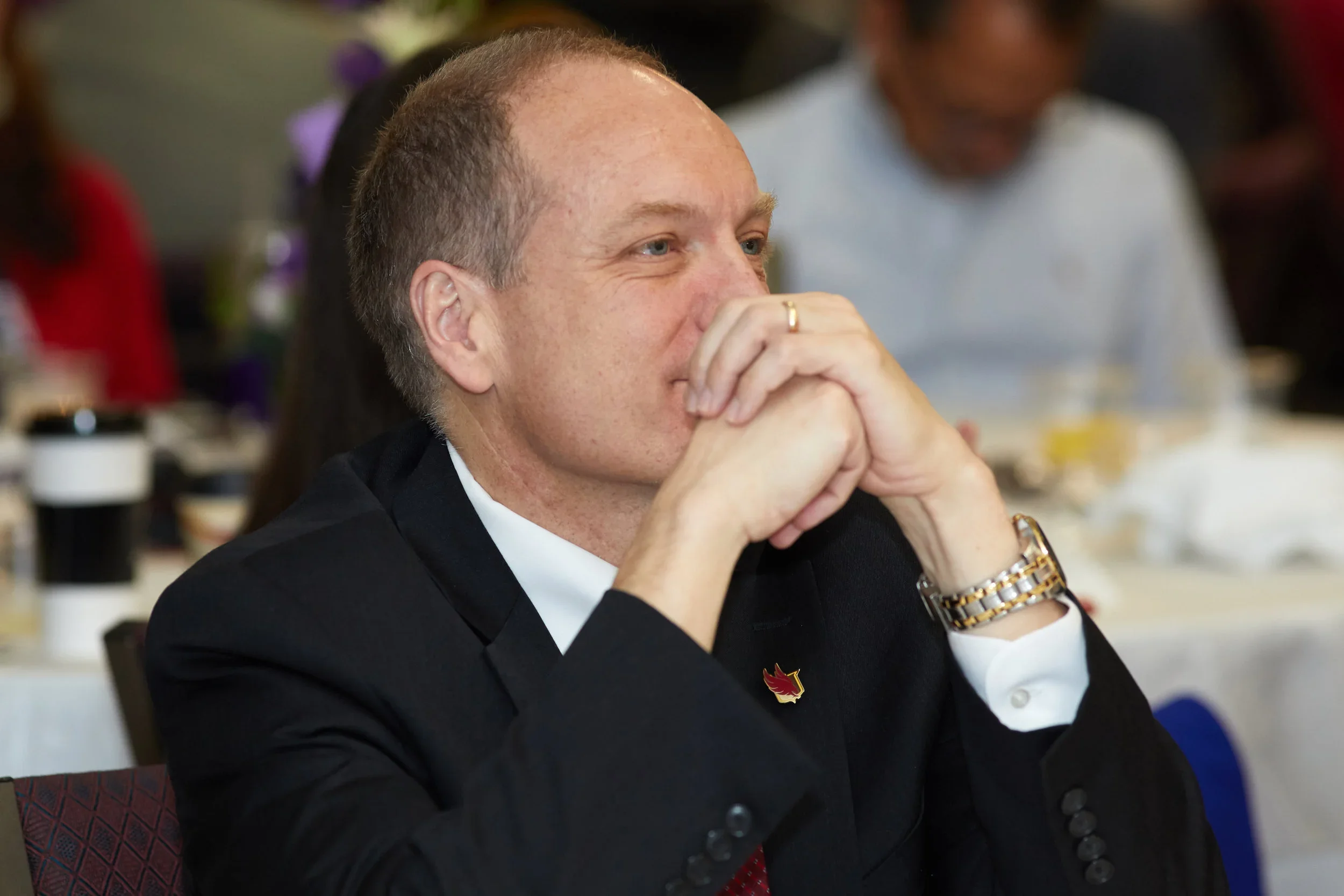 A man wearing a suit sitting with his elbows on a table and his hands folded by this mouth, appears to be at a banquet. 