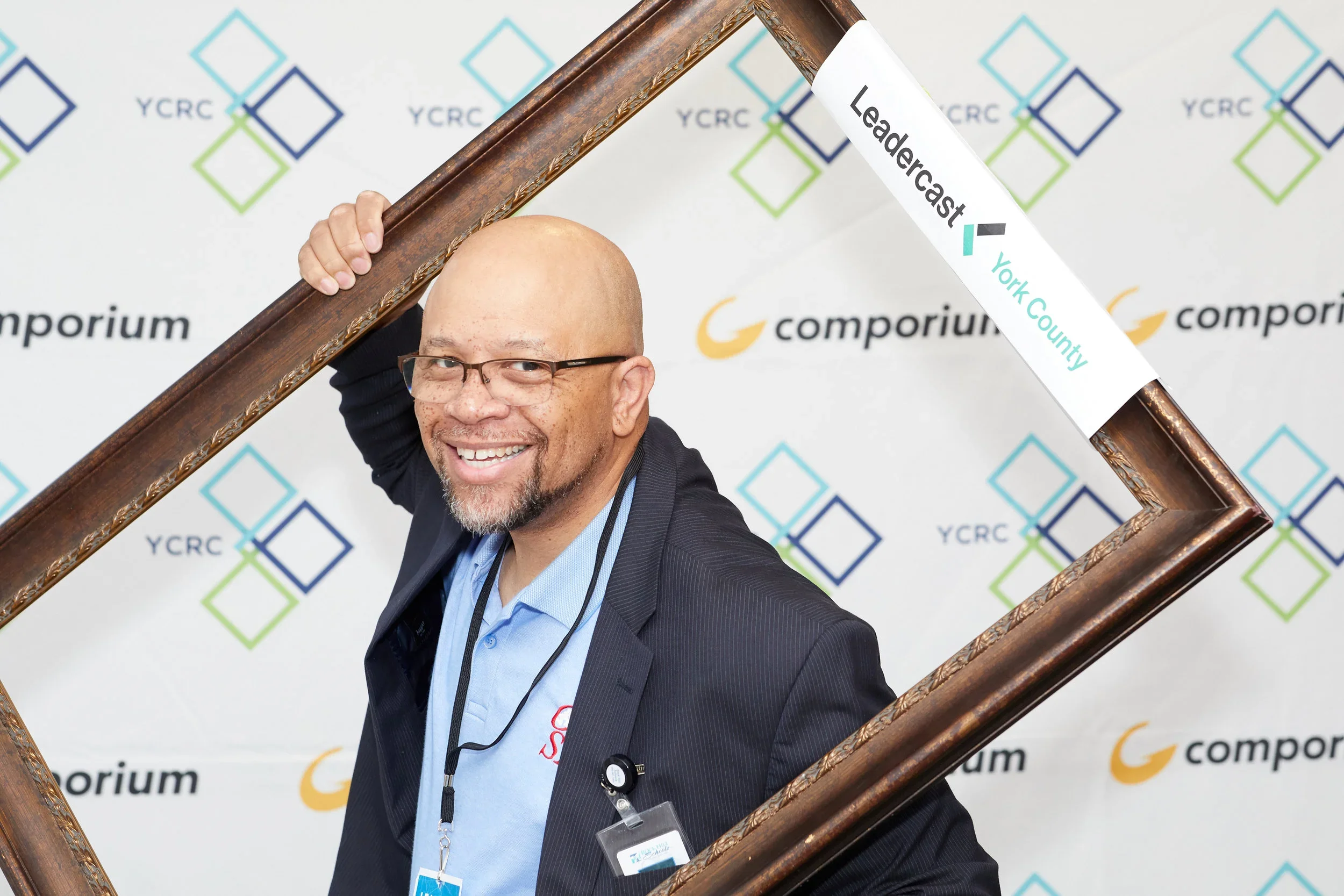A smiling man with glasses and a goatee wearing a suit jacket and a light blue dress shirt, holding a large ornate picture frame over his shoulder at a conference or event with a YCRC backdrop.