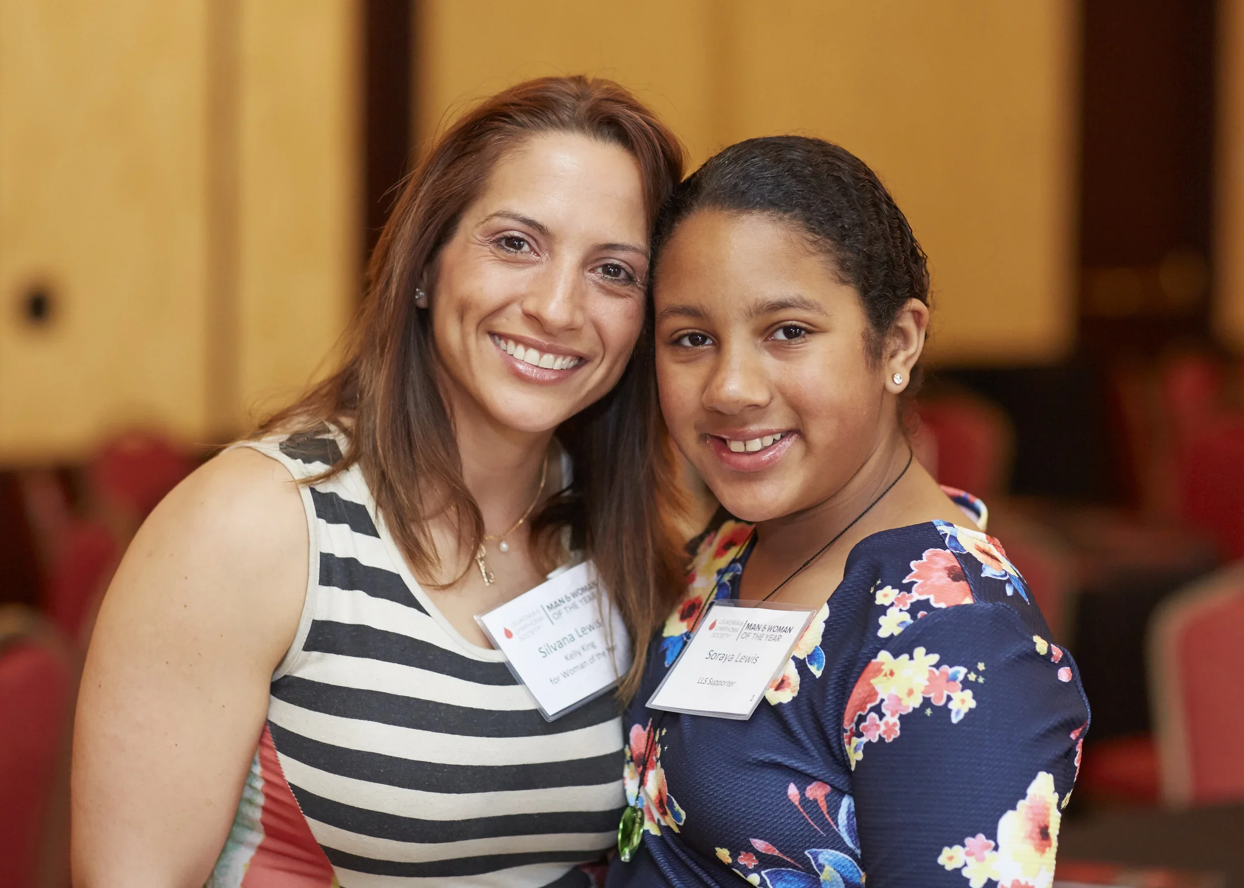 Two women smiling and standing close together, wearing name tags, in an indoor setting with yellow walls and red chairs in the background.