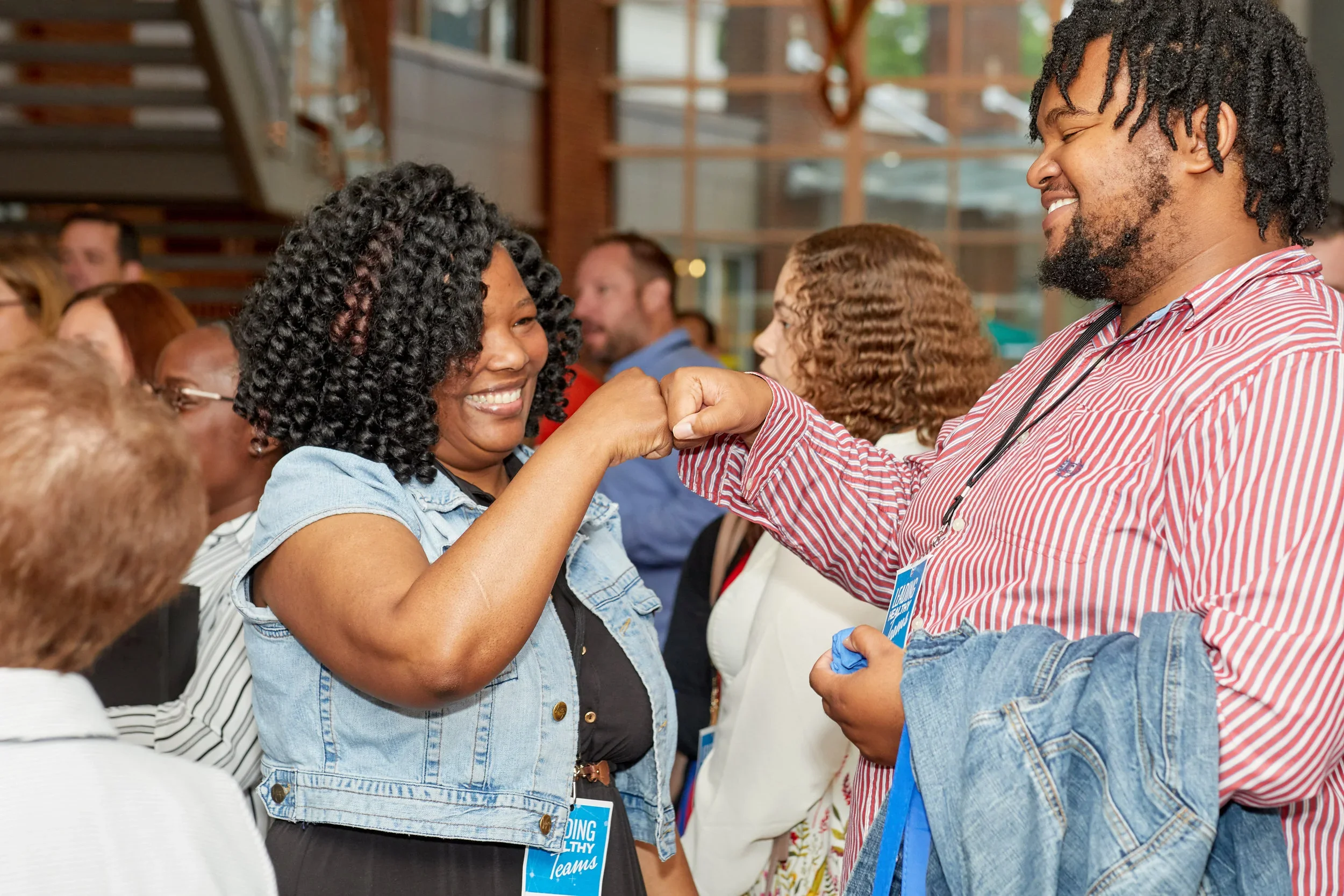 A woman and a man are fist-bumping and smiling at each other during a networking event, with other people in the background.