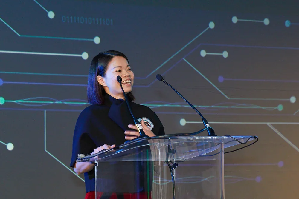 A woman speaking at a podium with a microphone, smiling, with a digital network and binary code background.