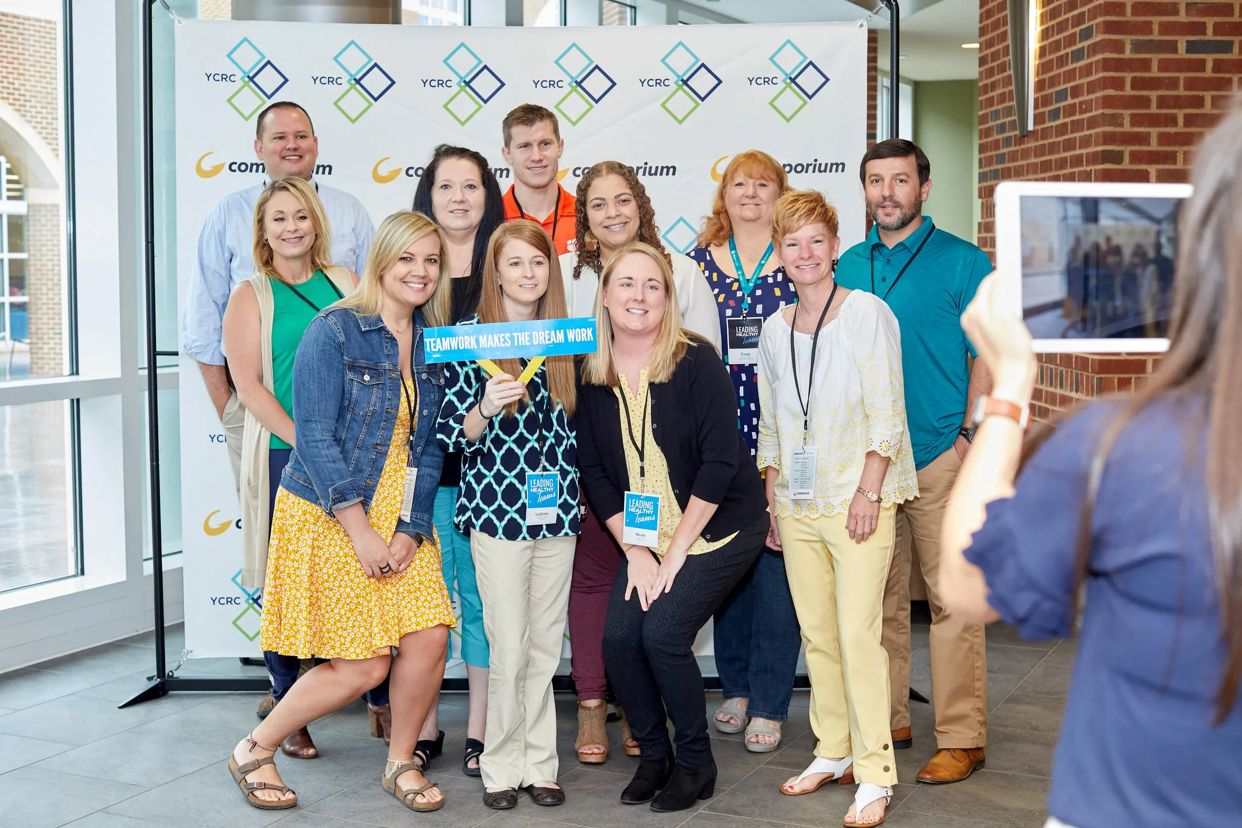 Group of people posing for a photo at a conference, with a woman holding a sign that says 'TEAMWORK MAKES THE DREAM WORK'. They are standing in front of a backdrop with 'YCRC' logos. One person is taking the photo with a tablet.