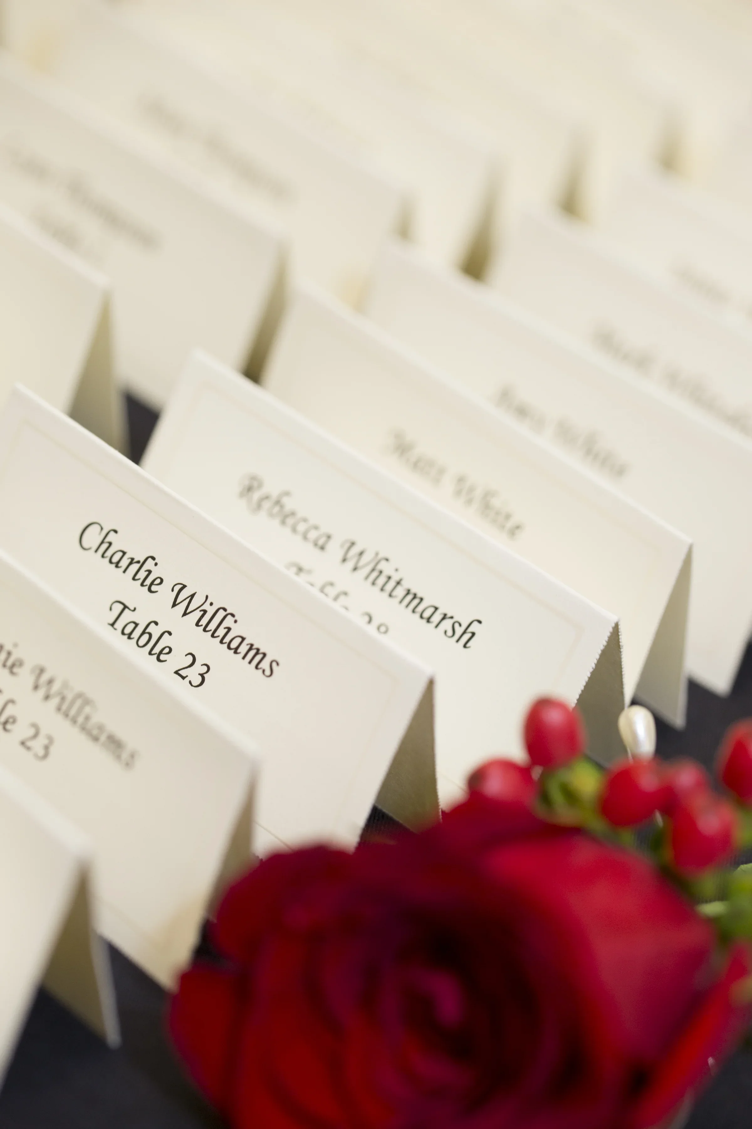 Wedding place cards with guest names, including Charlie Williams and Rebecca Whitmarsh, alongside a bouquet of red roses and berries.