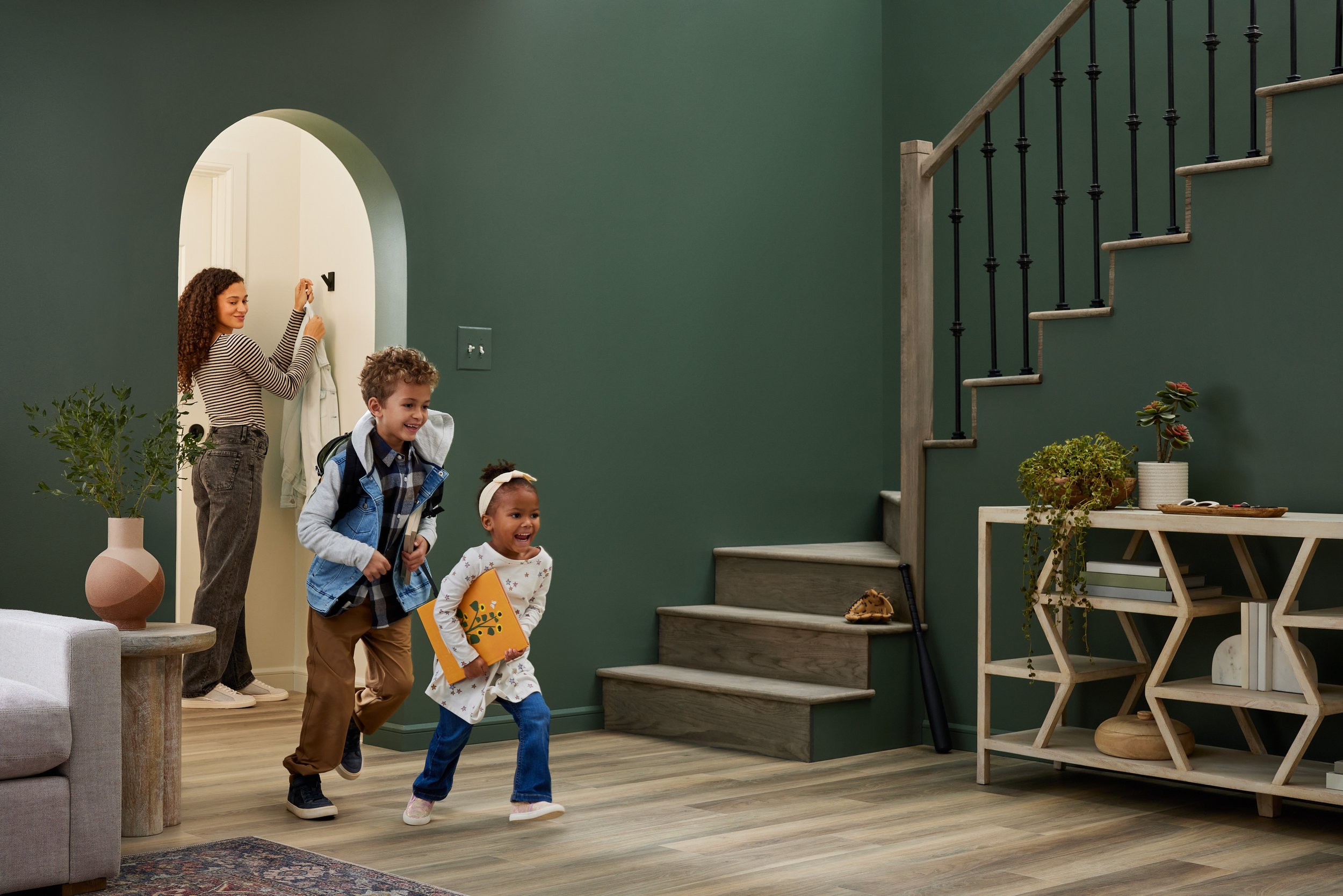 Two children run energetically in a living room while a woman hangs a coat on a hook. The room has a green wall, a staircase with a black railing, and a decorative shelf with plants and books. A baseball glove and bat are positioned on the stairs.