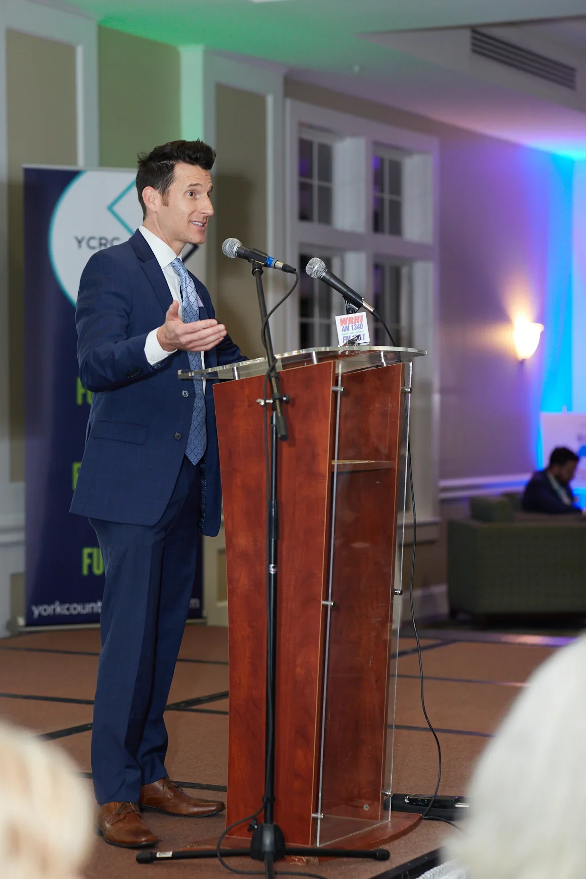 A man in a navy suit giving a speech at a podium during an event, with a banner and colorful lighting in the background.