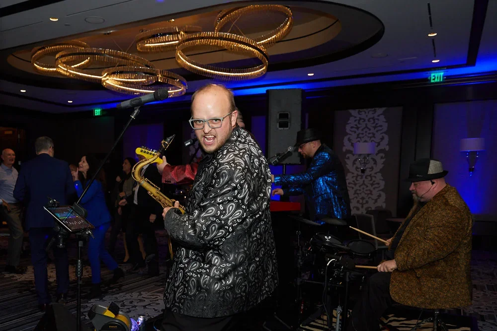 A band performing at an indoor event with a lively audience, featuring a saxophonist in the foreground expressing enthusiasm, with other musicians playing keyboard and drums, under decorative circular lighting fixtures.